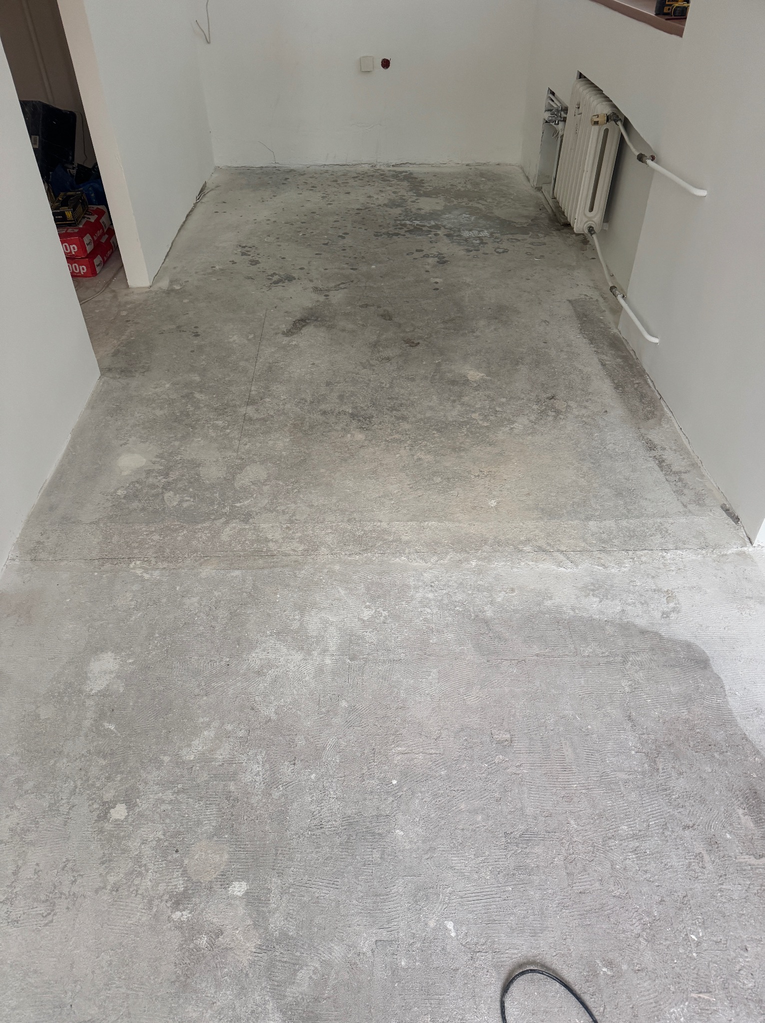 Przygotowana, szara, surowa podłoga w pustym pomieszczeniu z grzejnikiem, gotowa do dalszych prac wykończeniowych. Widoczne ślady po szlifowaniu.