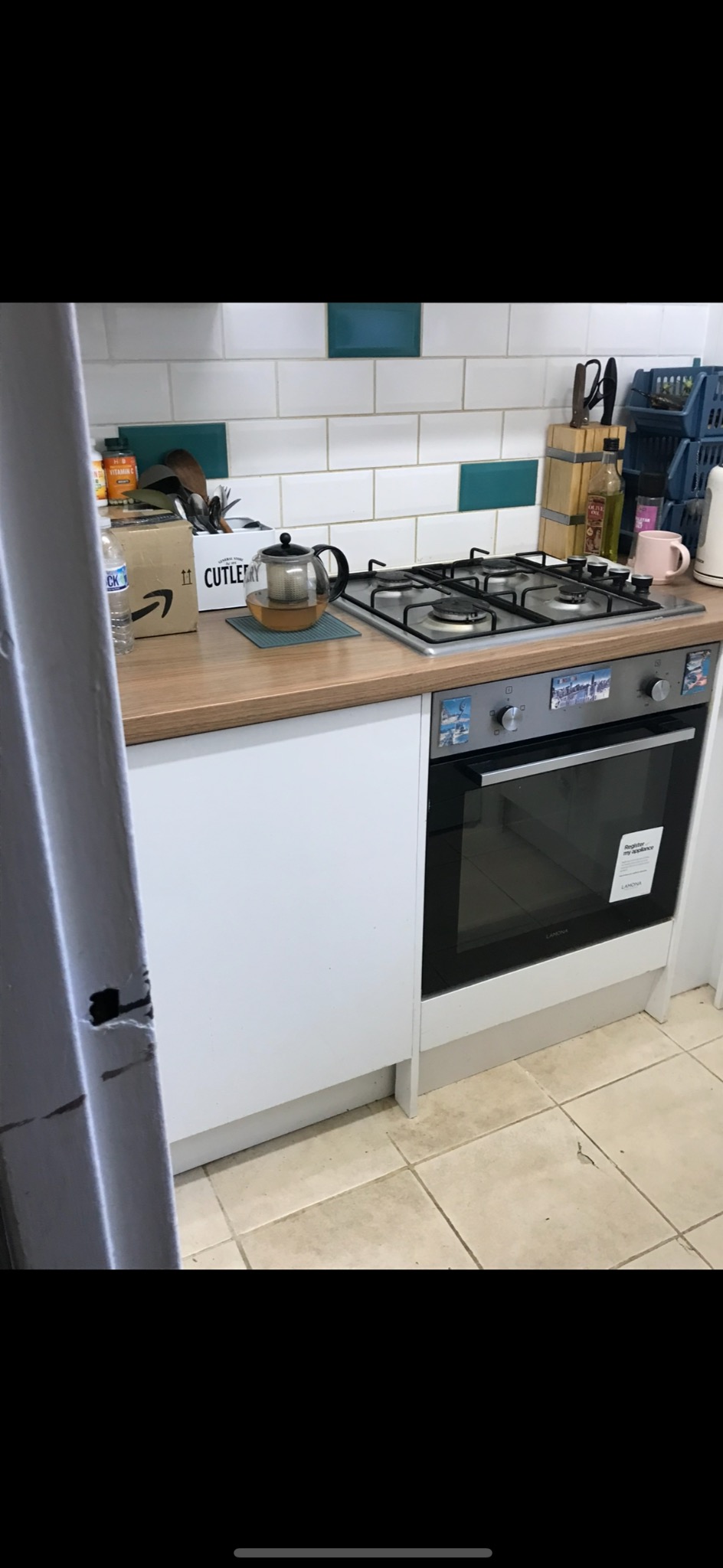 Jasna kuchnia z białymi szafkami, drewnianym blatem i płytą gazową. Na blacie czajnik, pojemnik na sztućce i pudełko. Białe płytki z turkusowymi akcentami na ścianie.