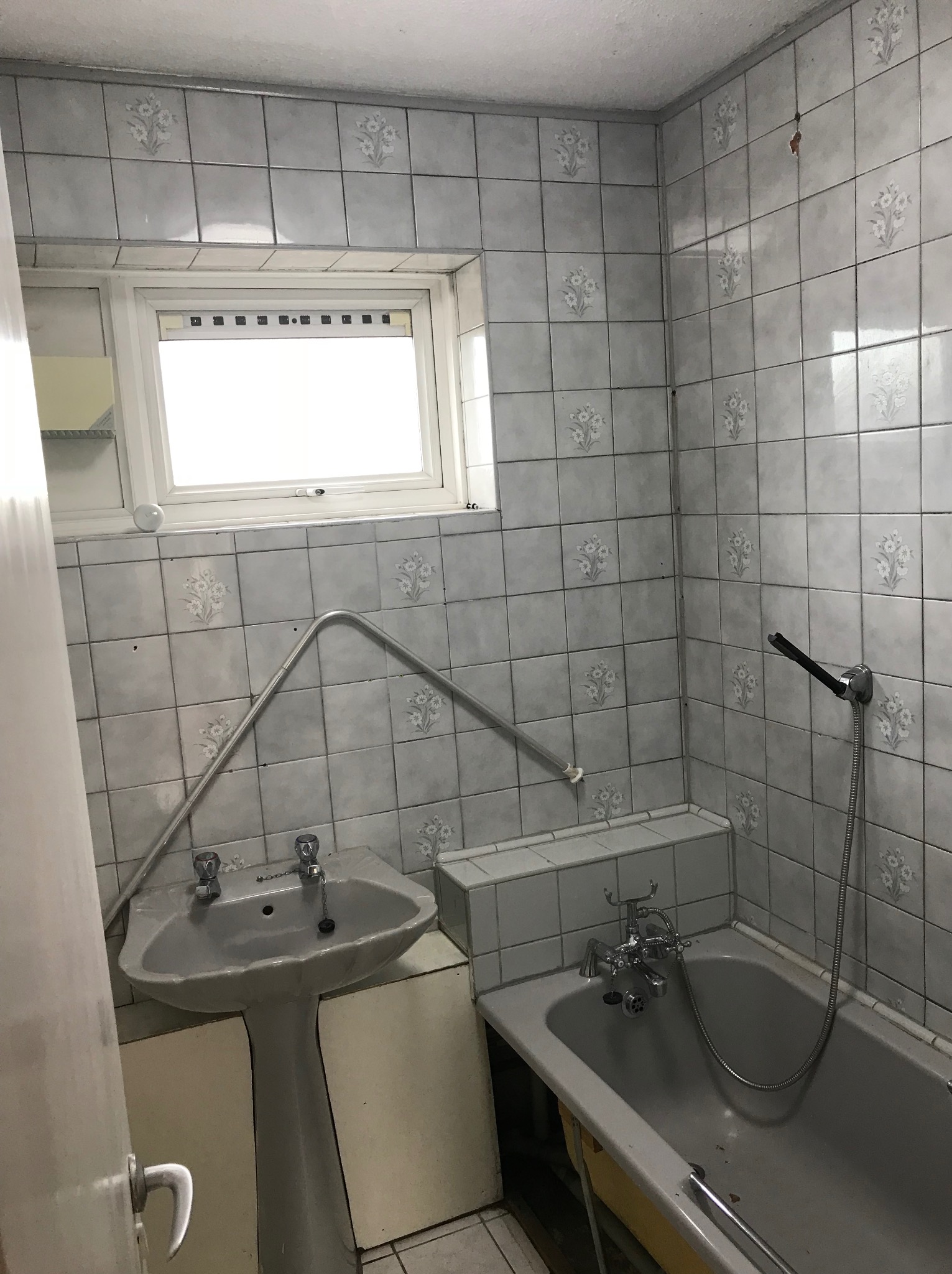 Łazienka przed remontem: szare płytki z kwiatowym wzorem, stara umywalka na postumencie, wanna z baterią. Widoczne ślady użytkowania i brak nowoczesnych rozwiązań.