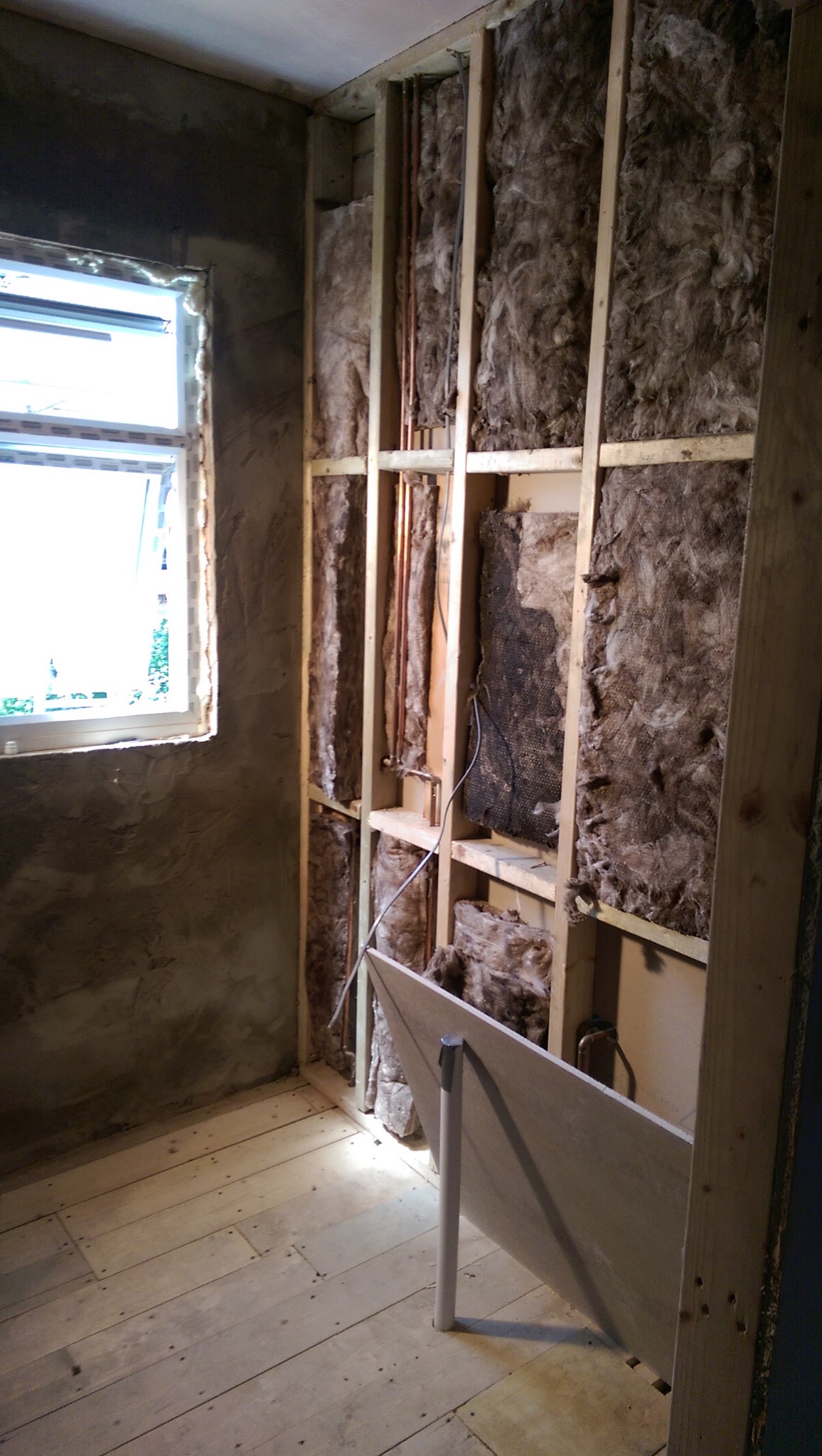 Ściana z izolacją z wełny mineralnej, drewnianą konstrukcją i instalacją hydrauliczną w trakcie prac wykończeniowych. Widoczna surowa podłoga i fragment płyty gipsowej.