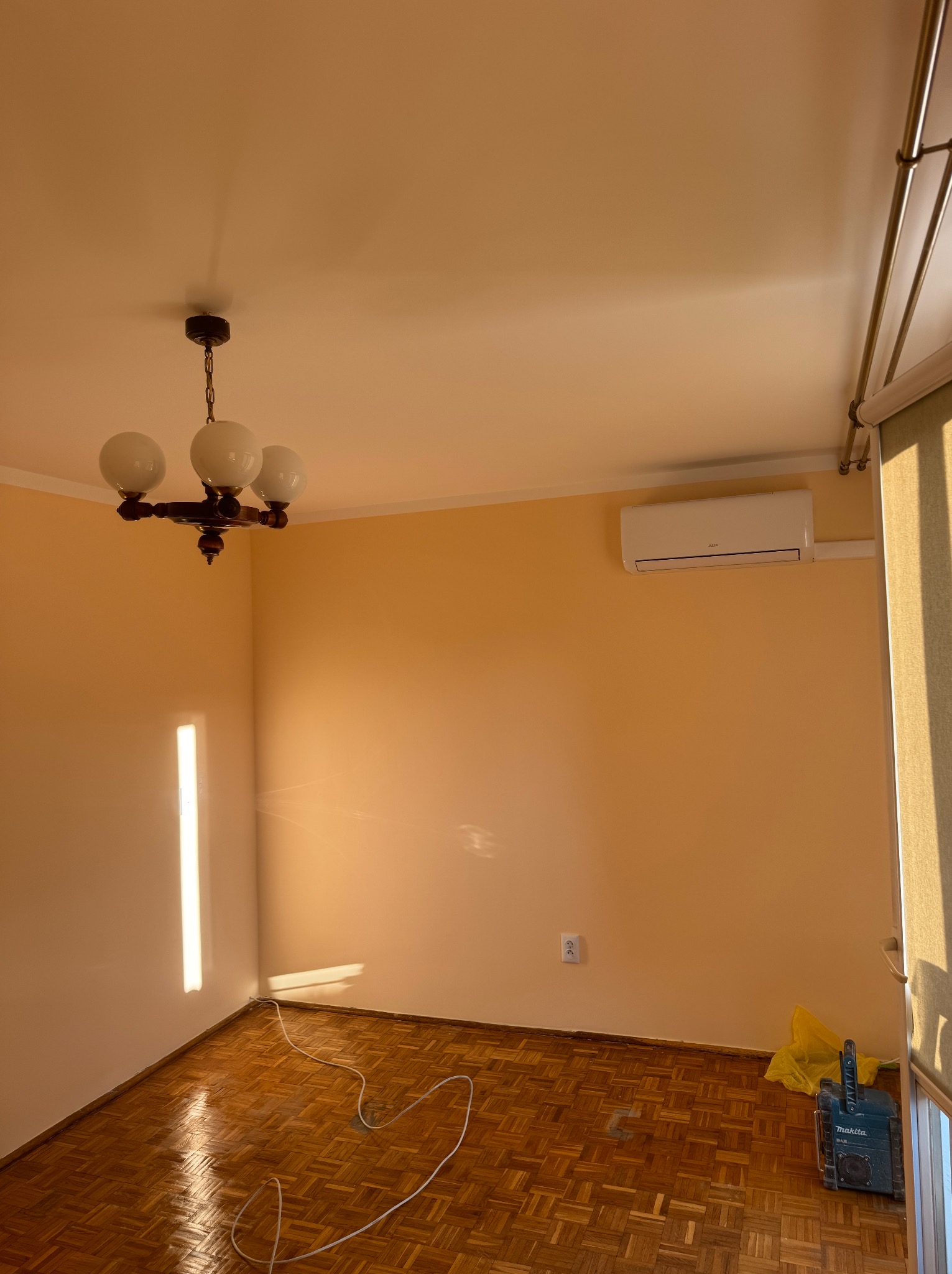 Pomarańczowy pokój po malowaniu z parkietem, lampą i klimatyzacją. Widoczny kabel i narzędzia, sugerujące prace wykończeniowe.