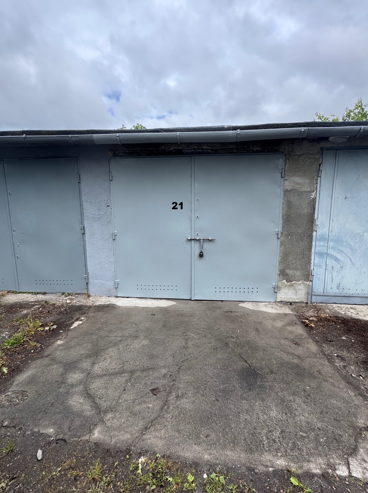 Szare, metalowe drzwi garażowe z numerem 21, zabezpieczone kłódką. Widoczne ślady użytkowania i betonowe elementy konstrukcyjne. Pochmurne niebo w tle.