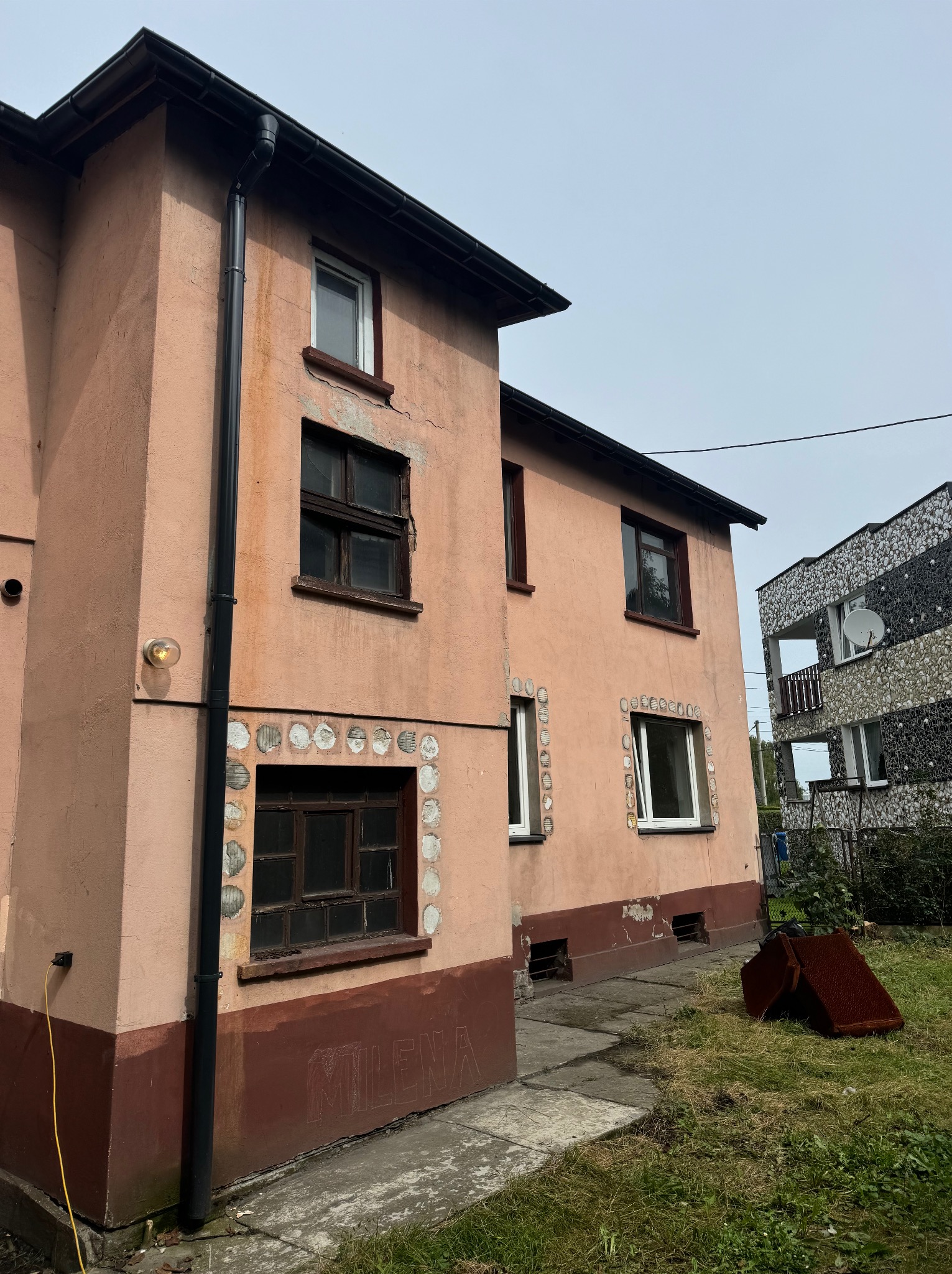 Dwupiętrowy budynek z różową elewacją, ciemnymi obramowaniami okien i dekoracyjnymi elementami wokół okien. Widoczne ślady użytkowania i napis 'MILENA' na cokole.