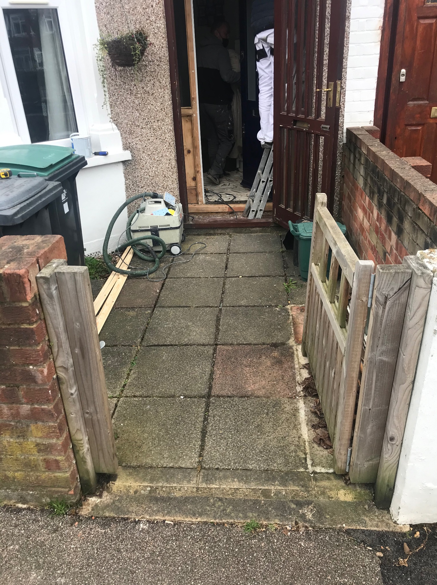 Remont wejścia do domu: malowanie drzwi wejściowych, widoczny odkurzacz przemysłowy, drabina i fragment drewnianego ogrodzenia. Prace w toku.