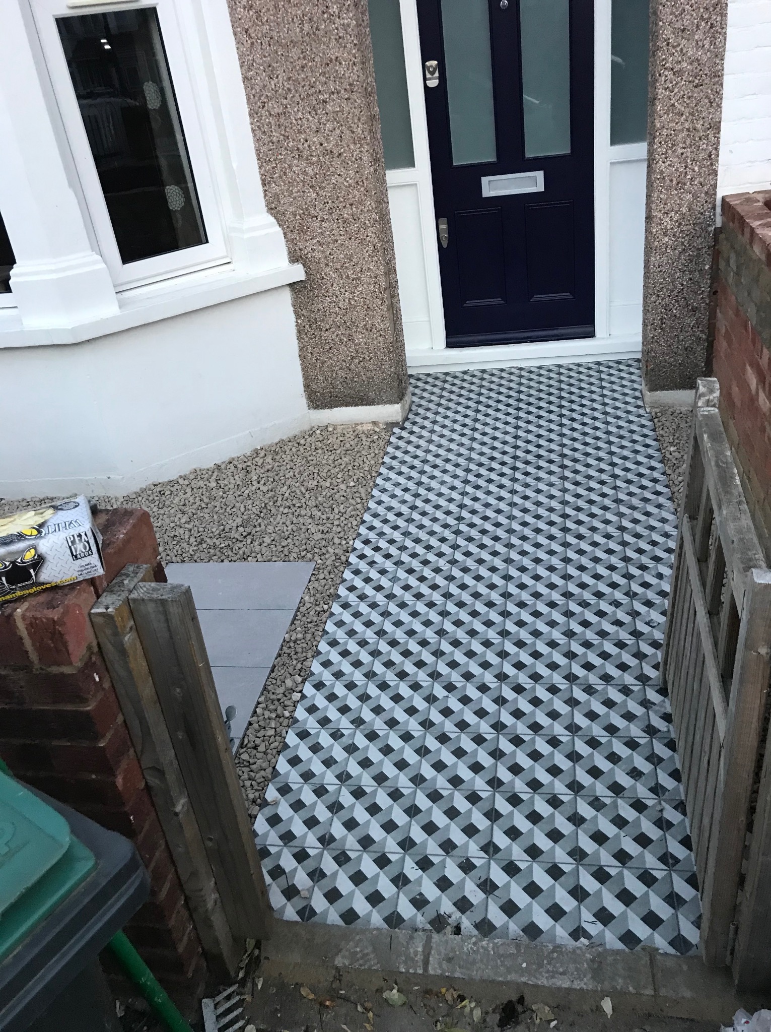 Ułożona geometryczna posadzka na zewnątrz budynku, prowadząca do ciemnych drzwi wejściowych. Widoczne kamyki, fragment okna i ceglany murek.