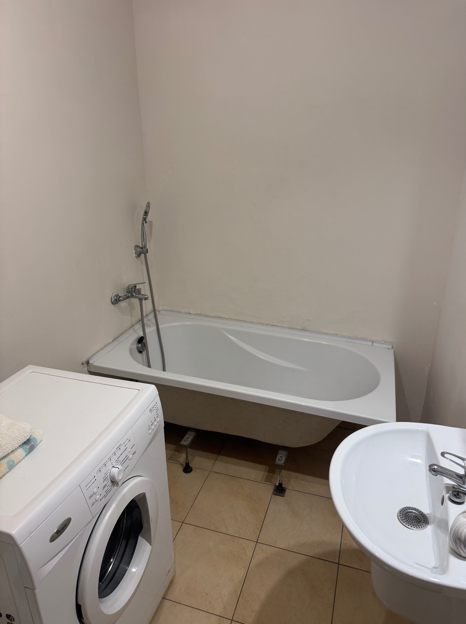 Jasna łazienka z białą wanną na nóżkach, pralką i umywalką. Beżowe płytki podłogowe. Widoczne ślady użytkowania na ścianach.