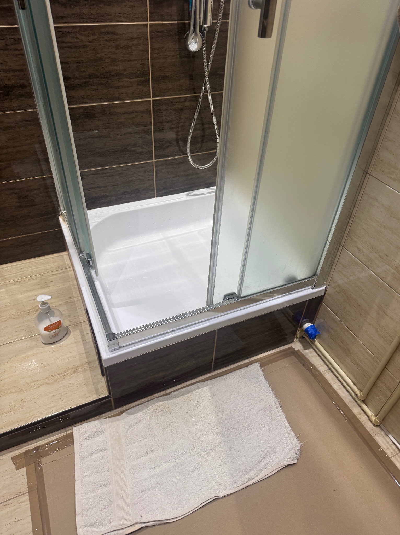 Biała wanna z rozsuwaną, częściowo otwartą kabiną prysznicową, brązowe i beżowe płytki na ścianach, ręcznik na podłodze. Widok z góry, oświetlenie dzienne.