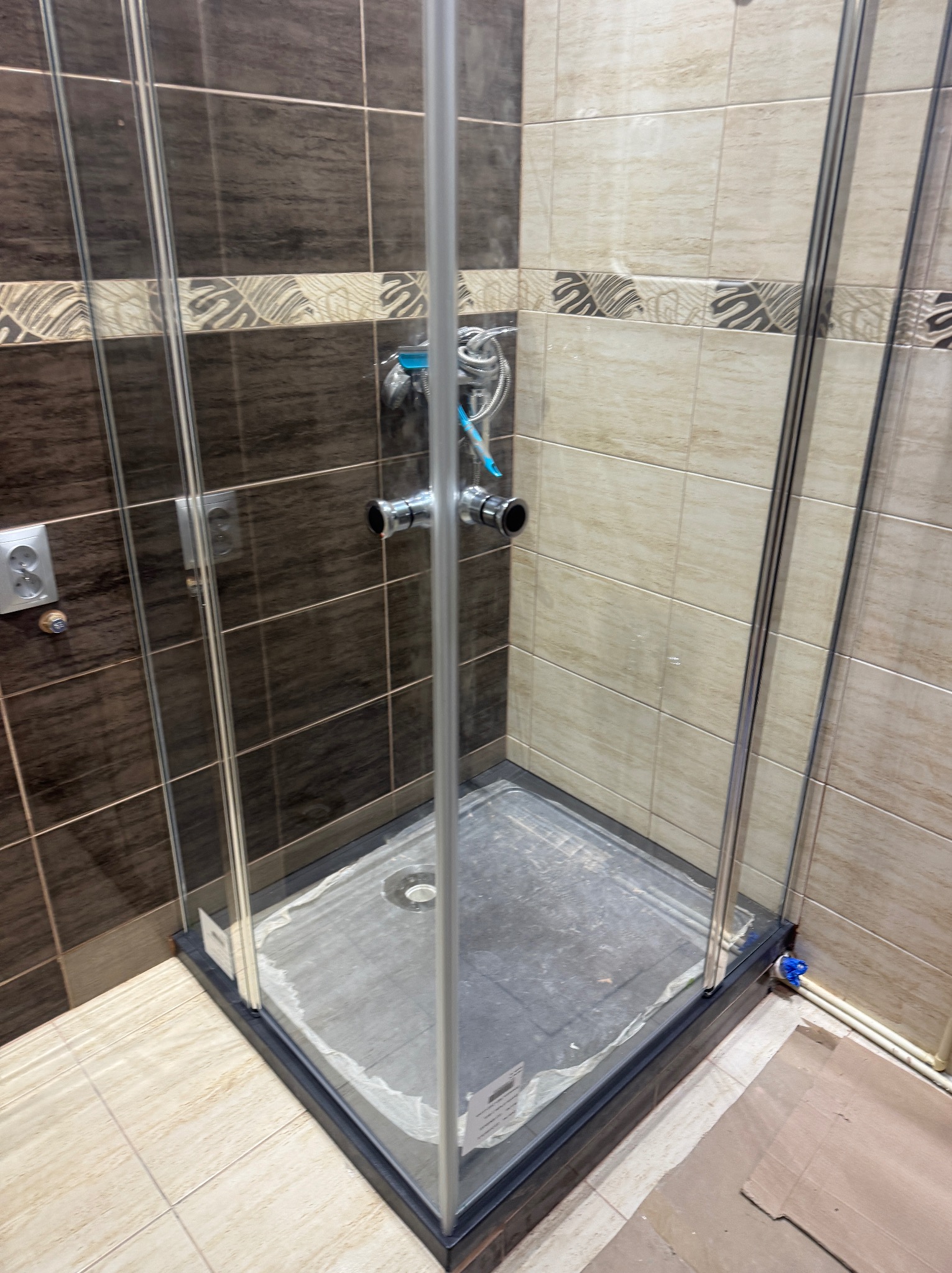 Kabina prysznicowa z chromowanymi profilami i dwukolorową glazurą w łazience. Widoczne przyłącza wodne i odpływ. Ułożone płytki podłogowe.