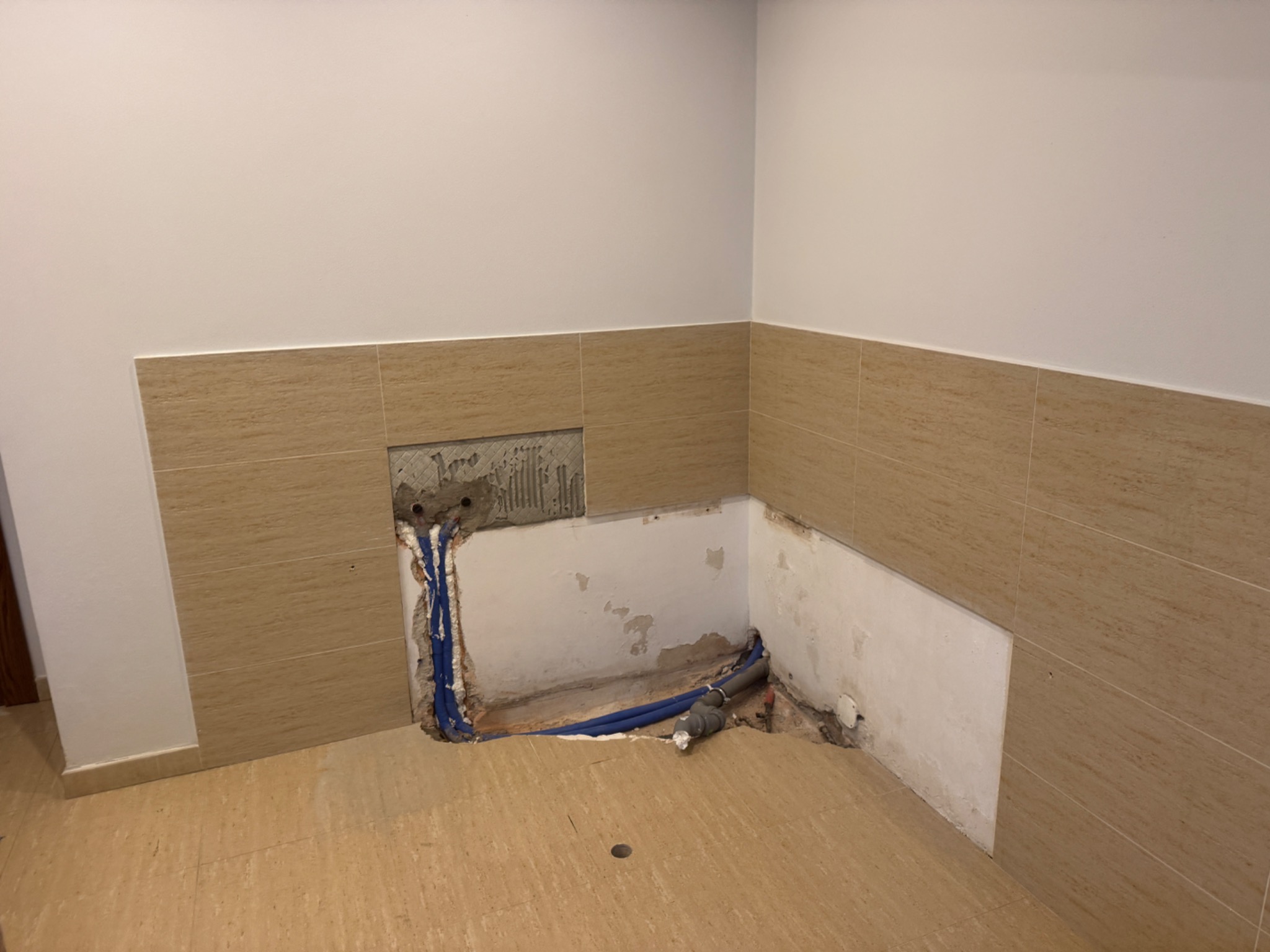 Fragment łazienki w trakcie prac: częściowo położone beżowe płytki, odsłonięte instalacje wodne i kanalizacyjne w ścianie, przygotowane do dalszych prac wykończeniowych.