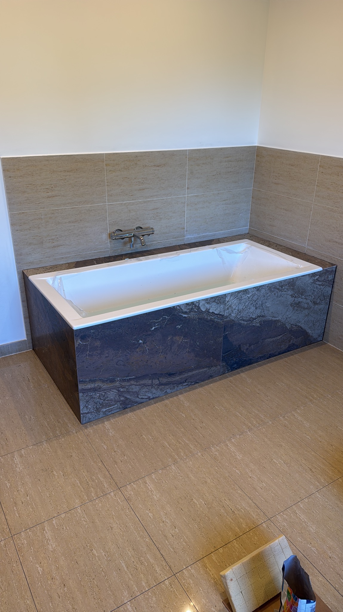 Nowoczesna łazienka z wanną obudowaną ciemnym marmurem, jasne płytki na ścianach i podłodze. Bateria wannowa chromowana. Wanna zabezpieczona folią ochronną.