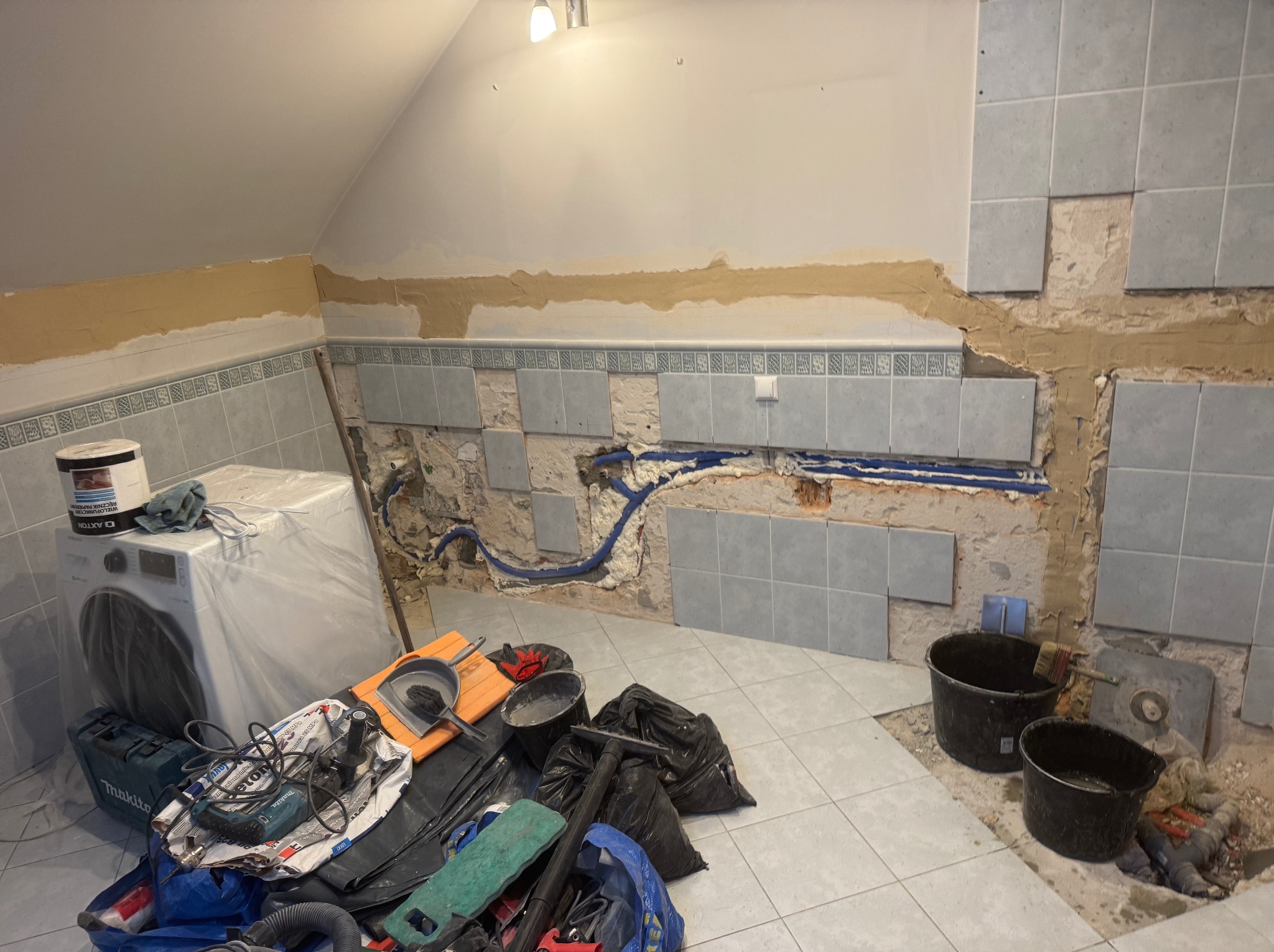 Fragment łazienki w trakcie remontu: częściowo położone szare płytki, widoczne instalacje, narzędzia i pralka zabezpieczona folią. Ujęcie z bliska, wnętrze pod skosem.