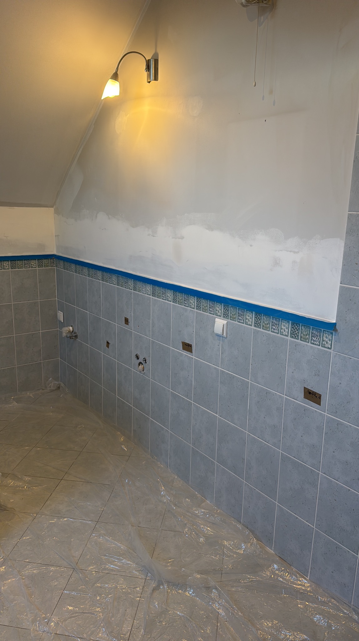 Łazienka w trakcie prac: szare płytki na ścianach do połowy wysokości, zabezpieczona folią podłoga, ściany przygotowane do malowania, widoczne punkty instalacji.