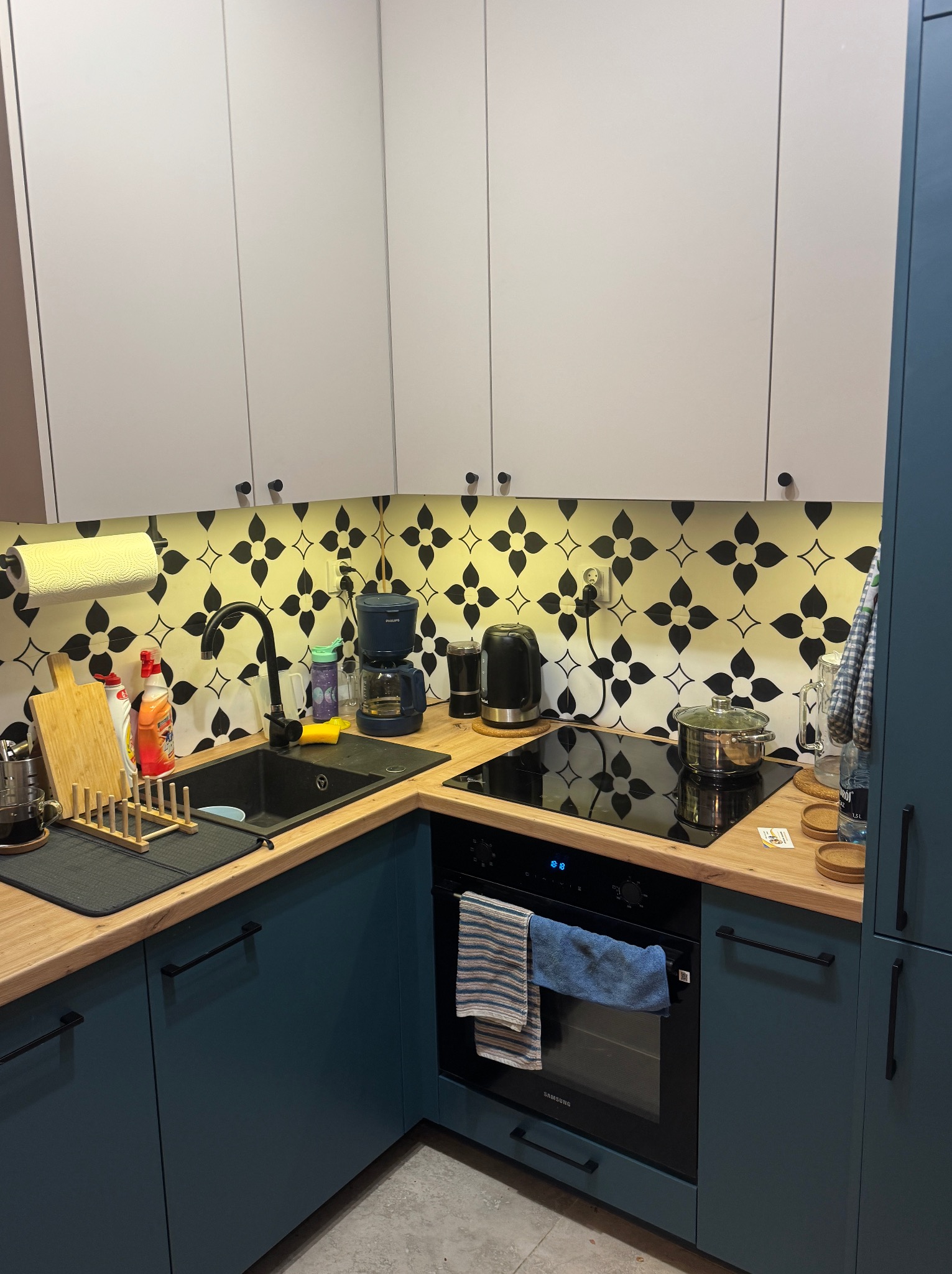 Nowoczesna kuchnia z białymi szafkami górnymi i niebieskimi dolnymi, drewnianym blatem i czarną armaturą. Na ścianie dekoracyjne płytki w kwiatowy wzór.