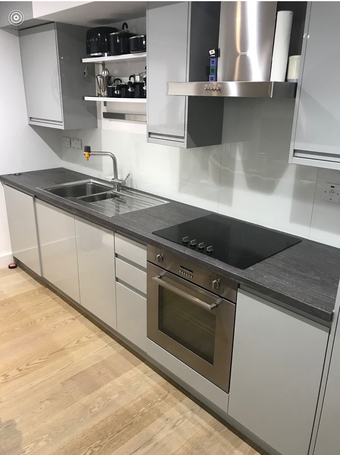 Nowoczesna kuchnia w odcieniach szarości z drewnianą podłogą. Widoczny zlew, płyta indukcyjna i piekarnik. Na półkach czarne garnki i akcesoria.