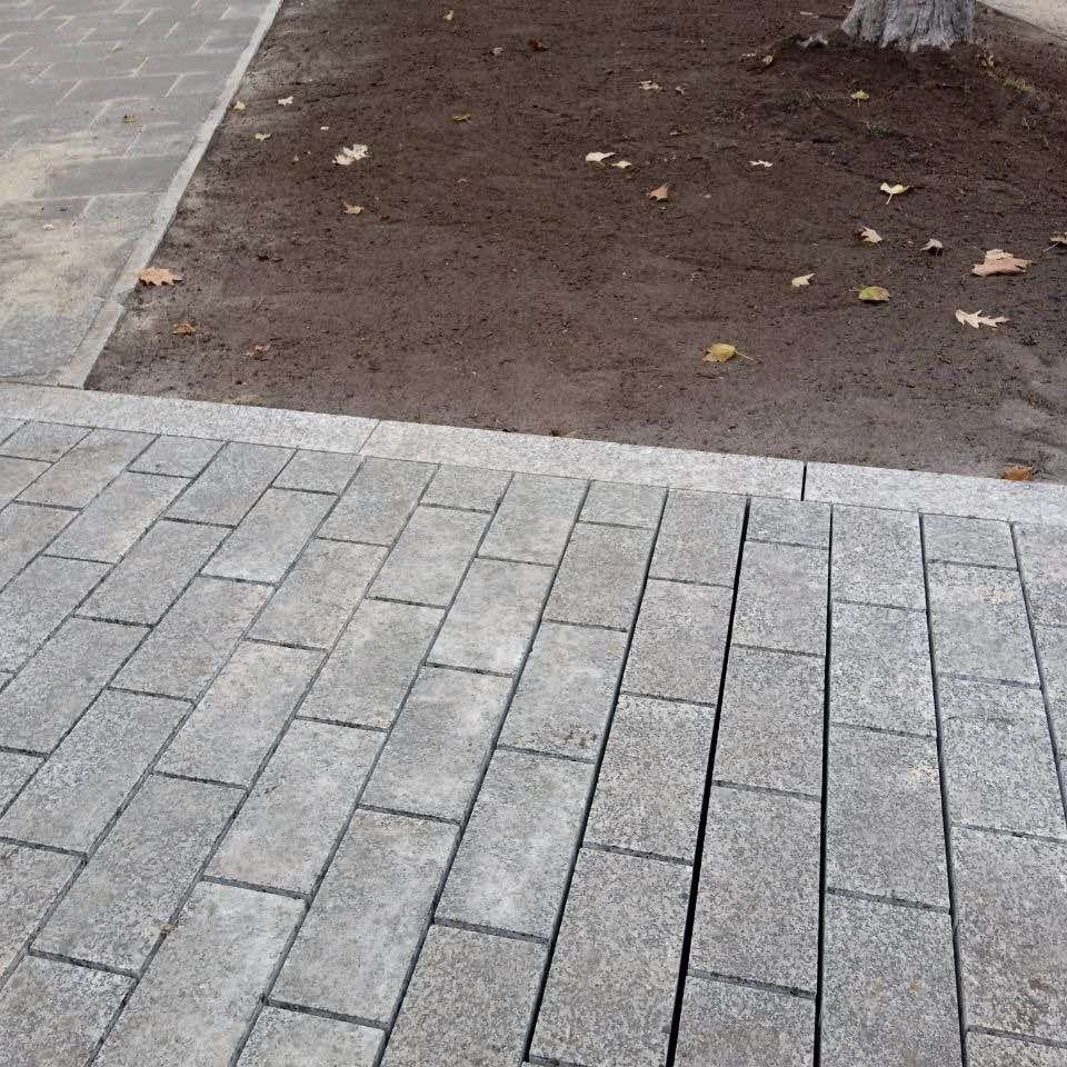 Szara kostka brukowa ułożona w regularny wzór, obok rabata z brązową ziemią i opadłymi liśćmi. Widok z góry, neutralne oświetlenie.