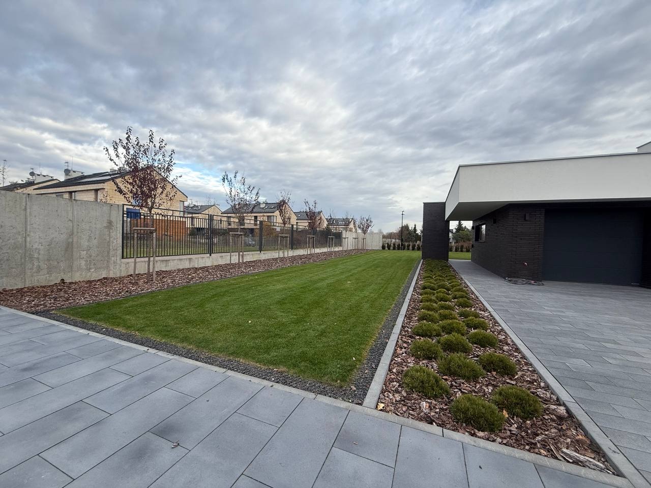 Nowoczesny podjazd z szarej kostki, obrzeża z grysu, trawnik i rabata z korą oraz niskimi krzewami. W tle dom z ciemną elewacją i szarym niebem.