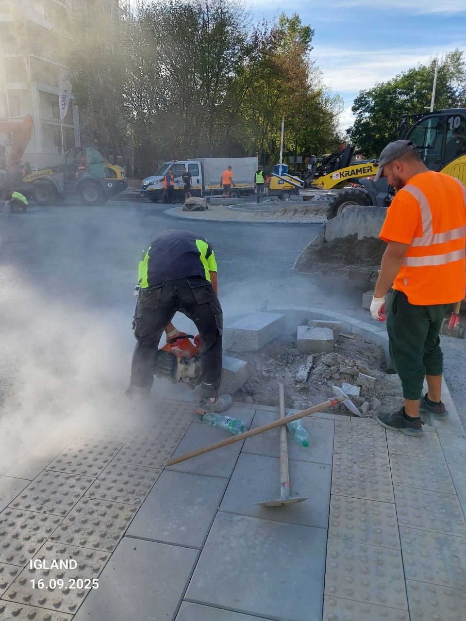 Pracownik tnie kostkę brukową na chodniku z wypustkami, obok leżą narzędzia i butelki wody. W tle maszyny budowlane i ekipa w odblaskowych kamizelkach.