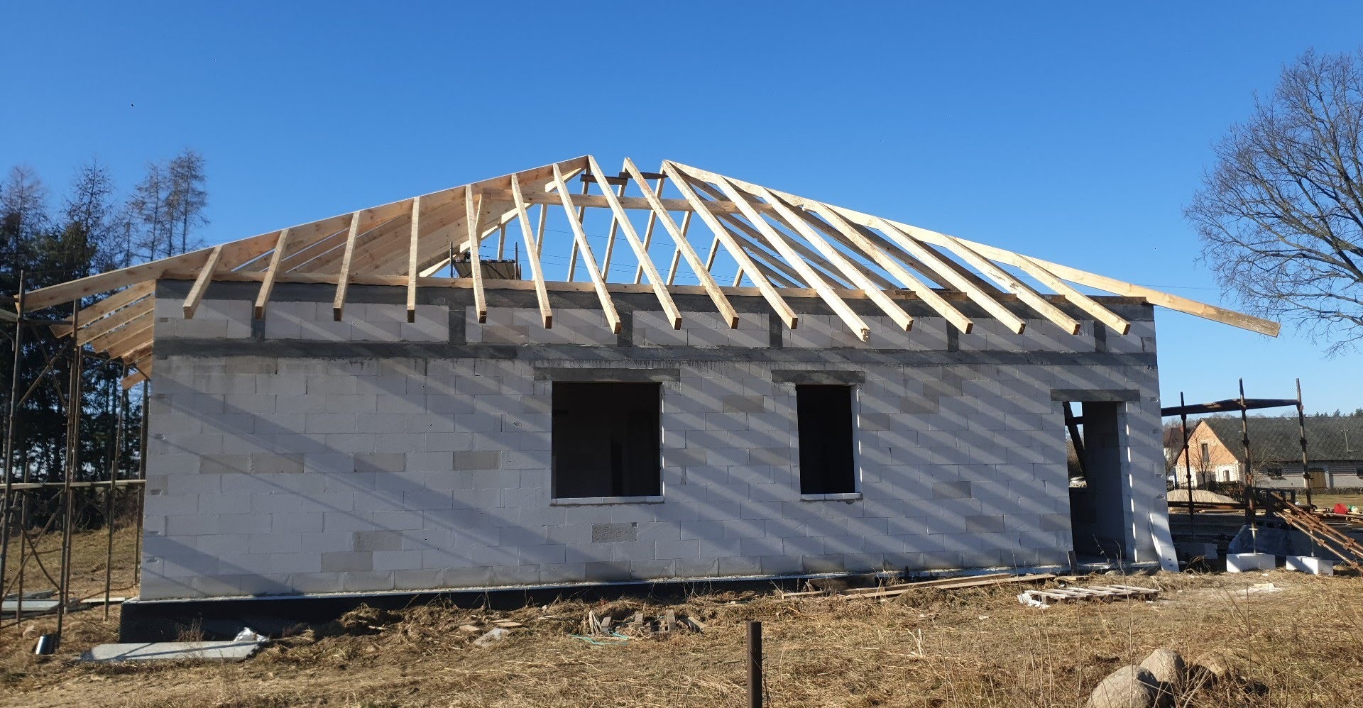 Budowa domu jednorodzinnego: ściany z betonu komórkowego i drewniana konstrukcja dachu w trakcie montażu, widok z zewnątrz w słoneczny dzień.