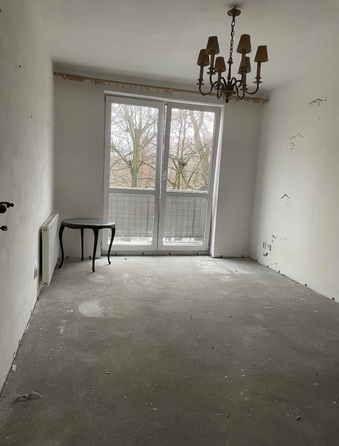 Pokój przed remontem w Warce: gołe ściany, surowa podłoga, okno z widokiem na drzewa, wiszący żyrandol. Przygotowanie do prac wykończeniowych.