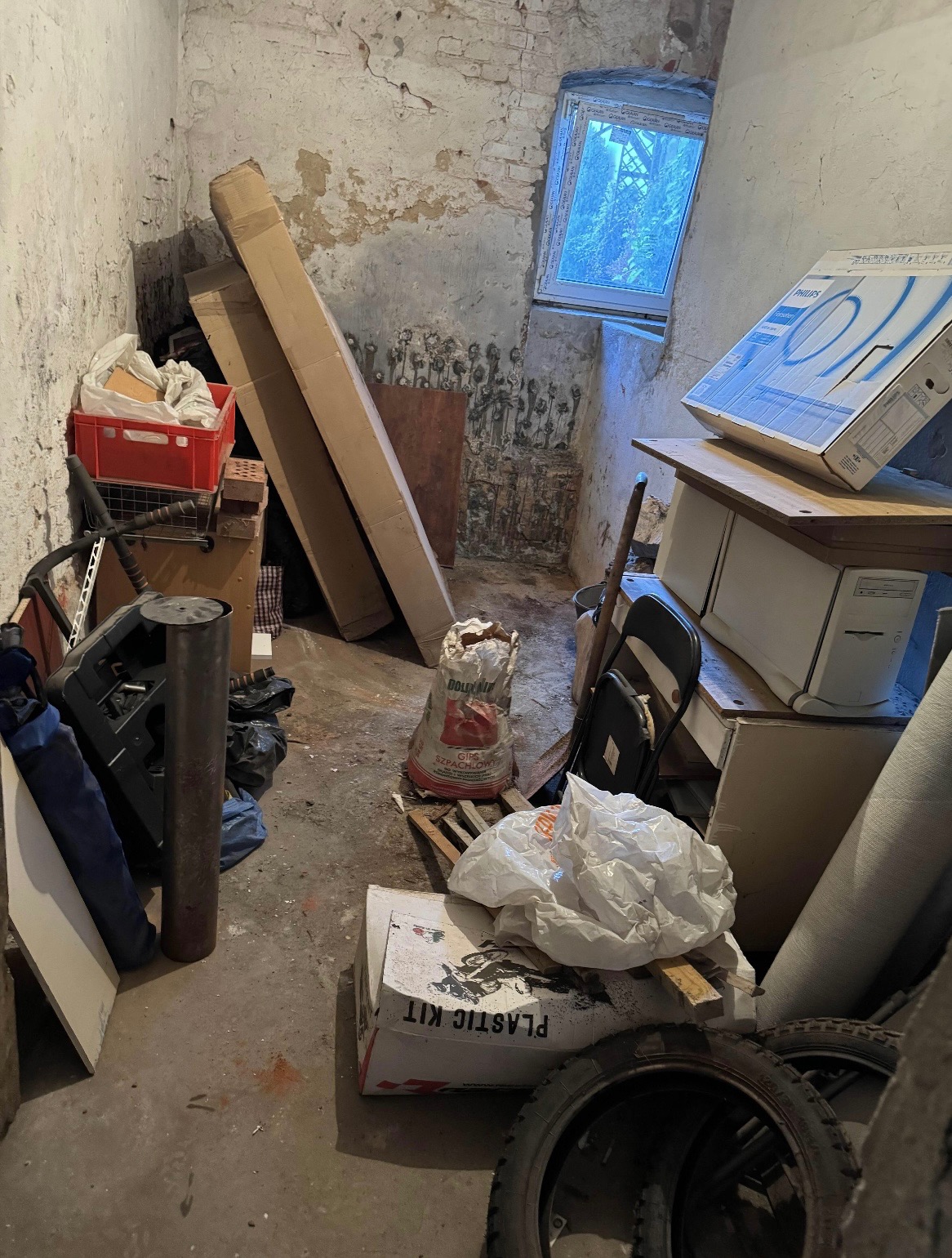 Zagracone pomieszczenie z kartonami, workami i starymi oponami. Widoczne ślady wilgoci na ścianach i małe okno w tle. Panuje ogólny nieporządek.
