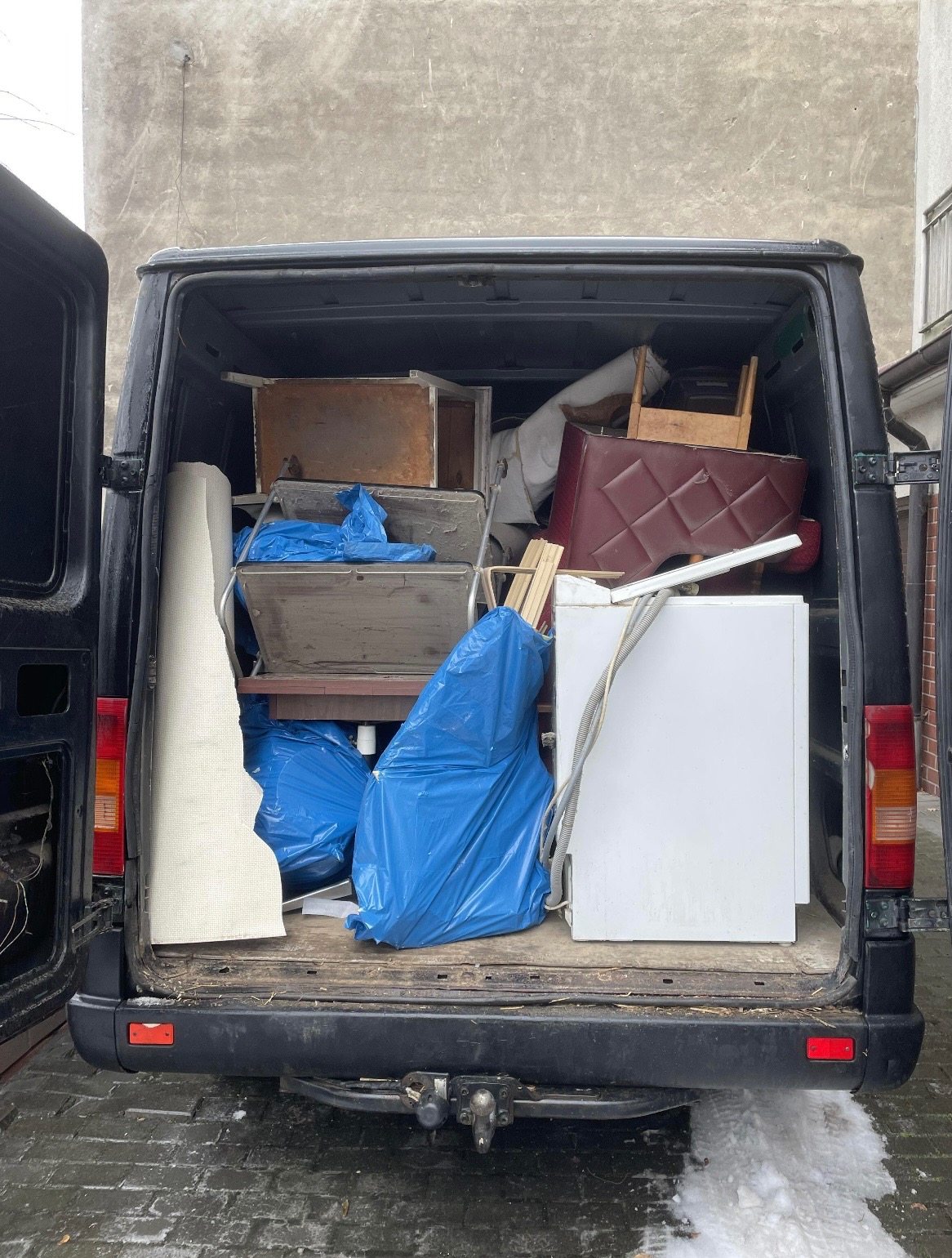 Czarny van z otwartymi drzwiami bagażnika, wypełniony meblami, sprzętem AGD i workami na śmieci. Widoczne elementy wyposażenia wnętrz i sprzęt gotowy do transportu.