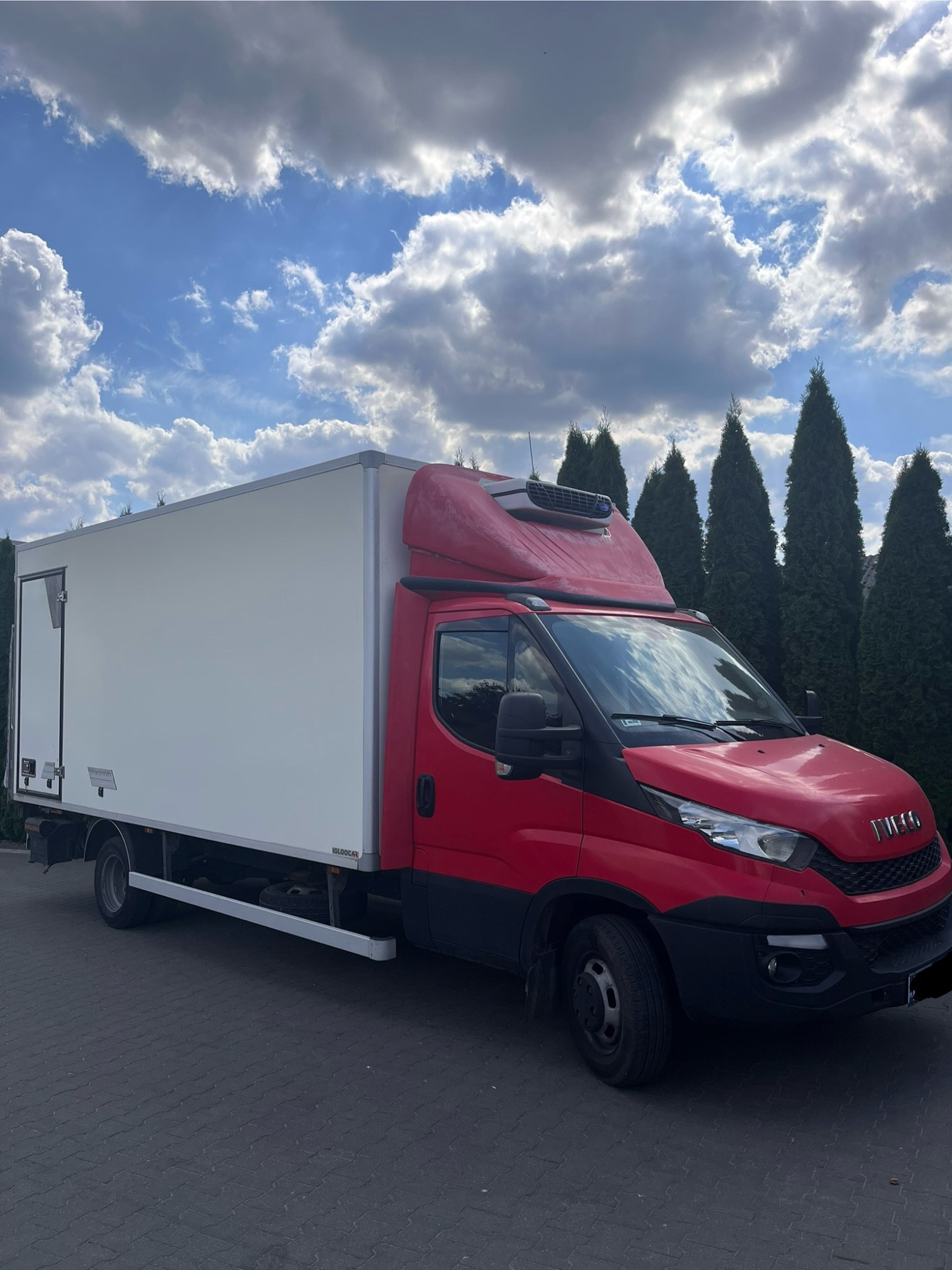 Czerwono-biała chłodnia Iveco na tle błękitnego nieba z obłokami i zielonych drzew. Pojazd zaparkowany na szarym bruku.