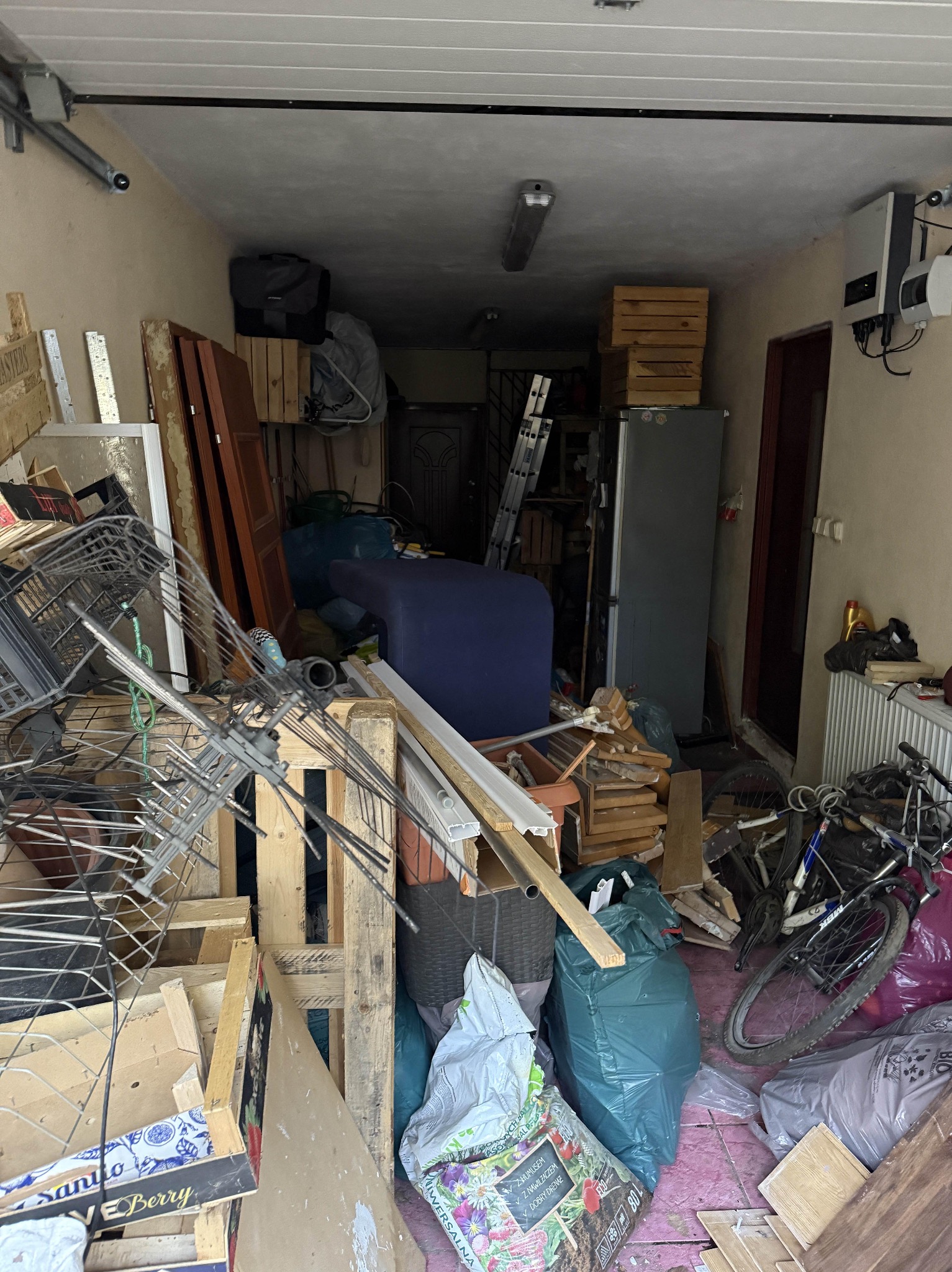 Zagracone pomieszczenie z meblami, sprzętami i odpadami, przygotowane do opróżnienia. Widoczne skrzynie, rower, worki i inne przedmioty w nieładzie.