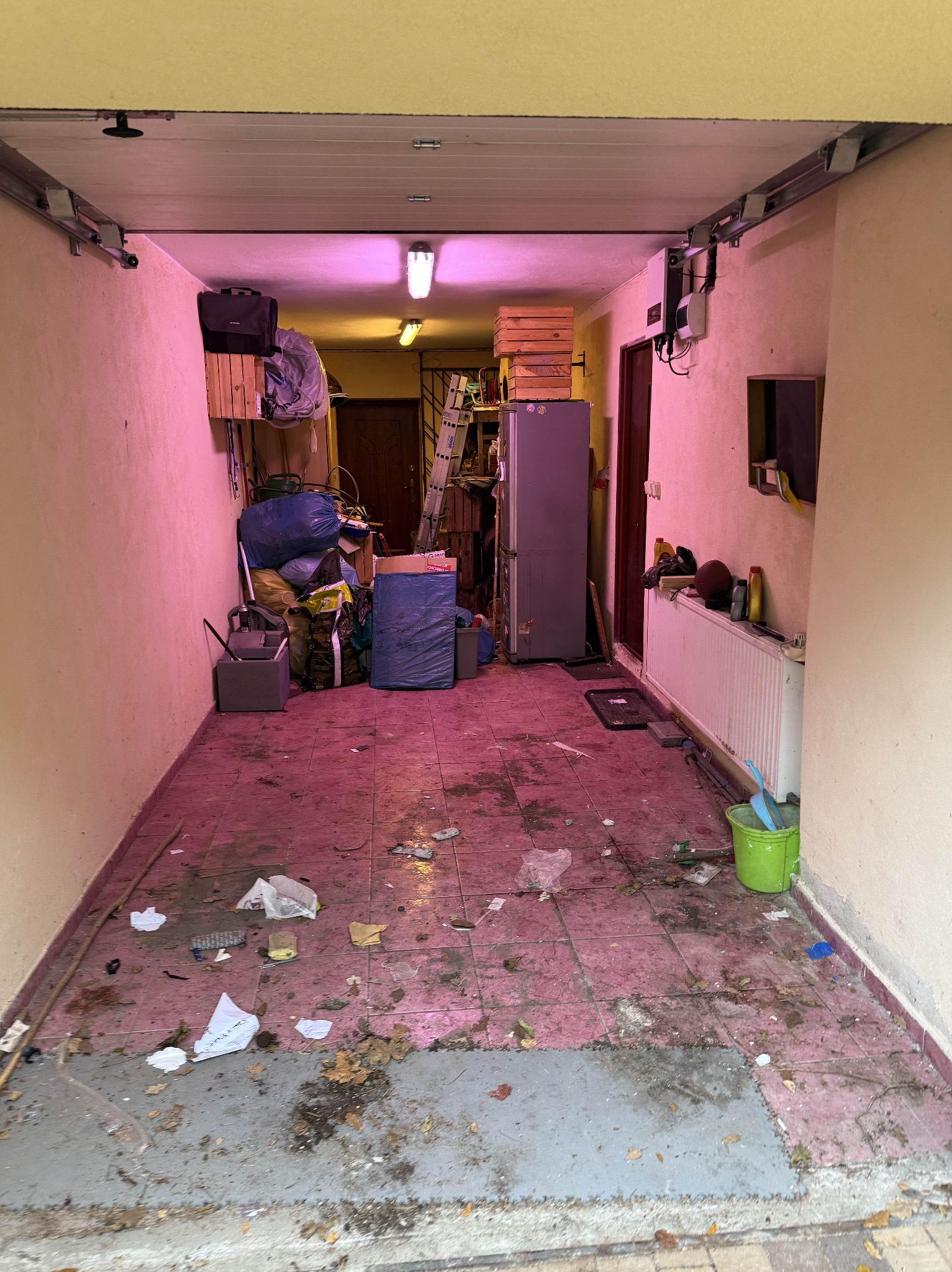 Zagracony garaż z różową poświatą: worki, kartony, sprzęt AGD, śmieci na podłodze z płytek. Widok na wnętrze z otwartą bramą garażową.