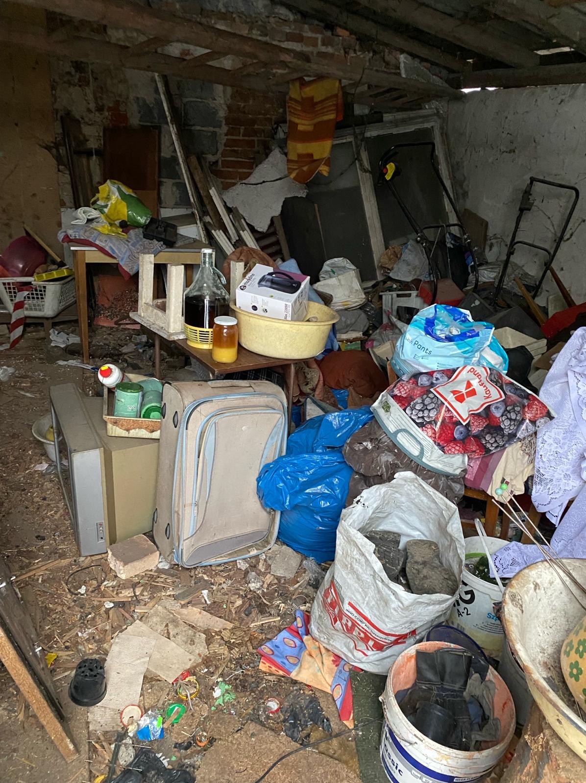 Zabałaganione pomieszczenie z różnymi przedmiotami: walizka, telewizor, butelki, torby z zakupami, zabawki i inne. Widoczne ślady zaniedbania i brak porządku.