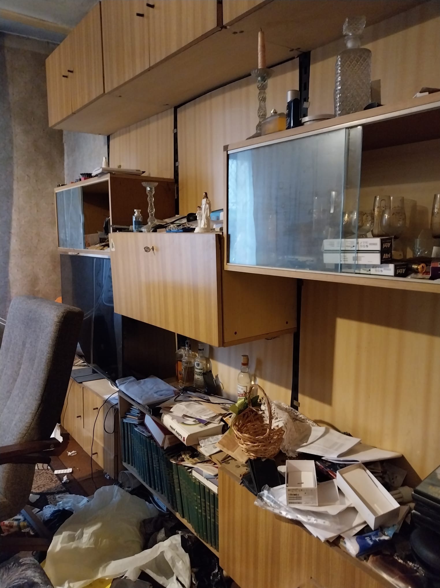 Zaniedbane wnętrze pokoju z meblościanką, na której panuje bałagan. Na podłodze widoczne śmieci i porozrzucane przedmioty. Krzesło częściowo zasłania widok.