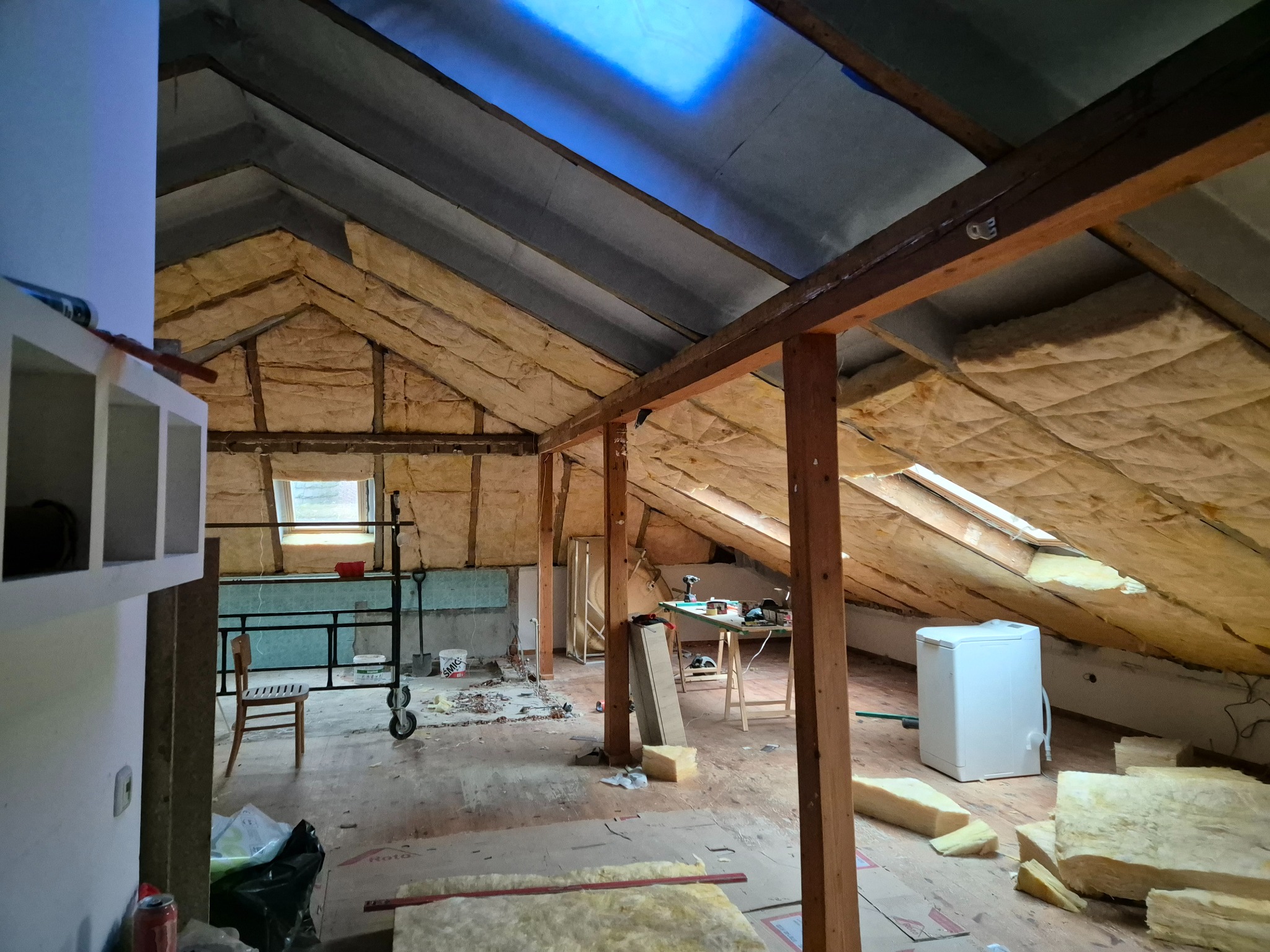 Ocieplenie poddasza wełną mineralną, widoczne drewniane belki konstrukcyjne, okno dachowe i narzędzia pracy. Prace wykończeniowe na poddaszu.