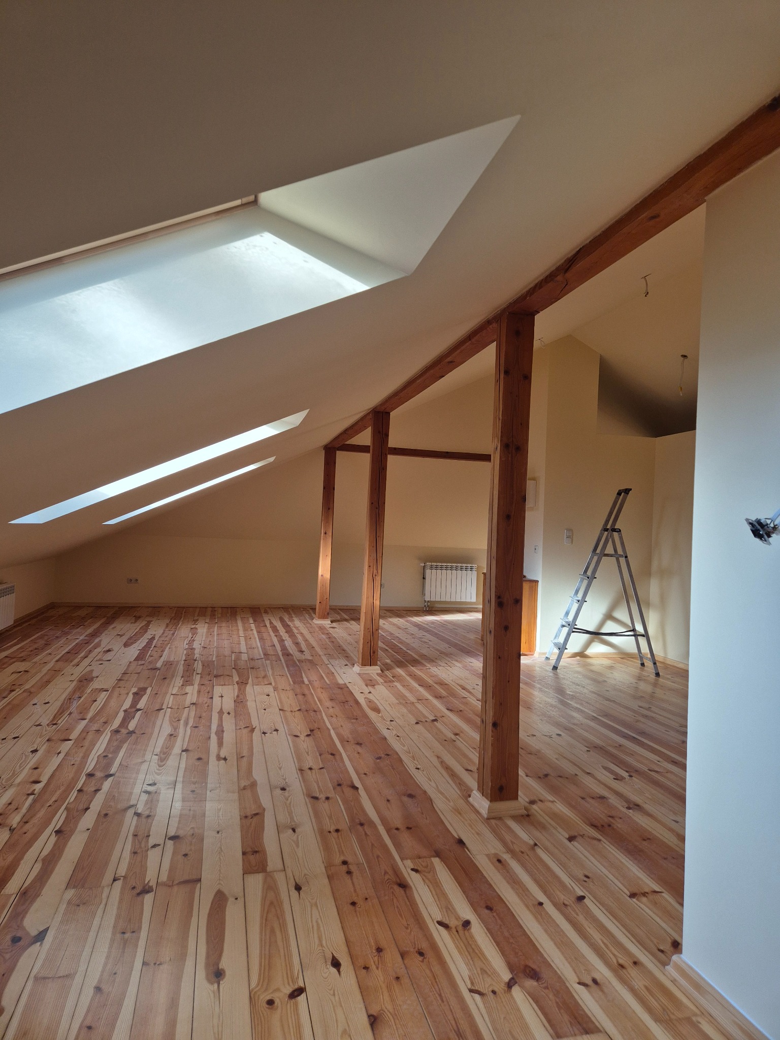 Jasne poddasze po remoncie: drewniana podłoga, belki konstrukcyjne, okna dachowe w skosie. Metalowa drabina oparta o ścianę. Widok na grzejnik w tle.