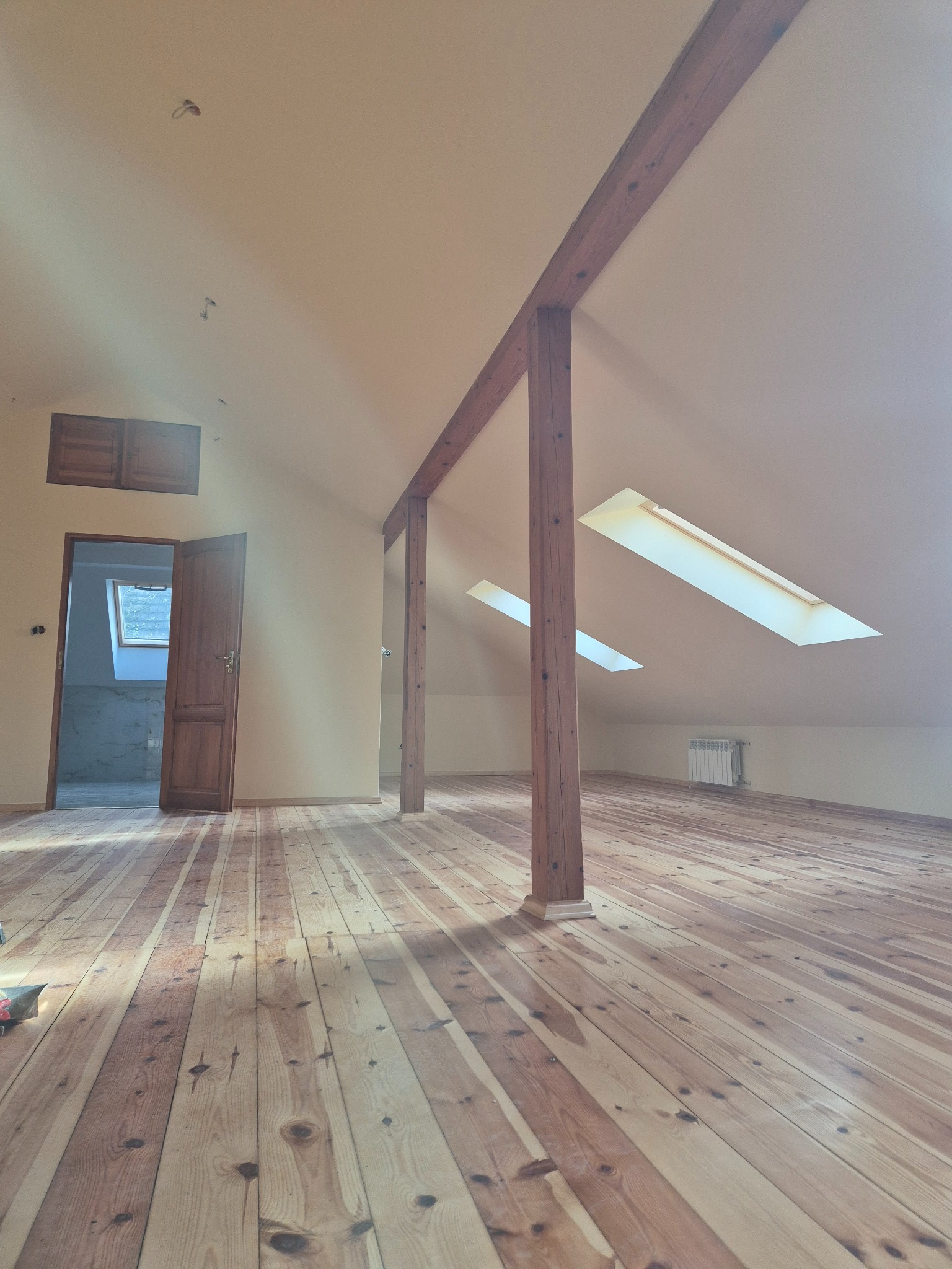 Jasne poddasze z drewnianą podłogą i belkami. Widoczne okna dachowe i grzejnik. Otwór drzwiowy prowadzi do kolejnego pomieszczenia. Świeżo położona podłoga z desek.
