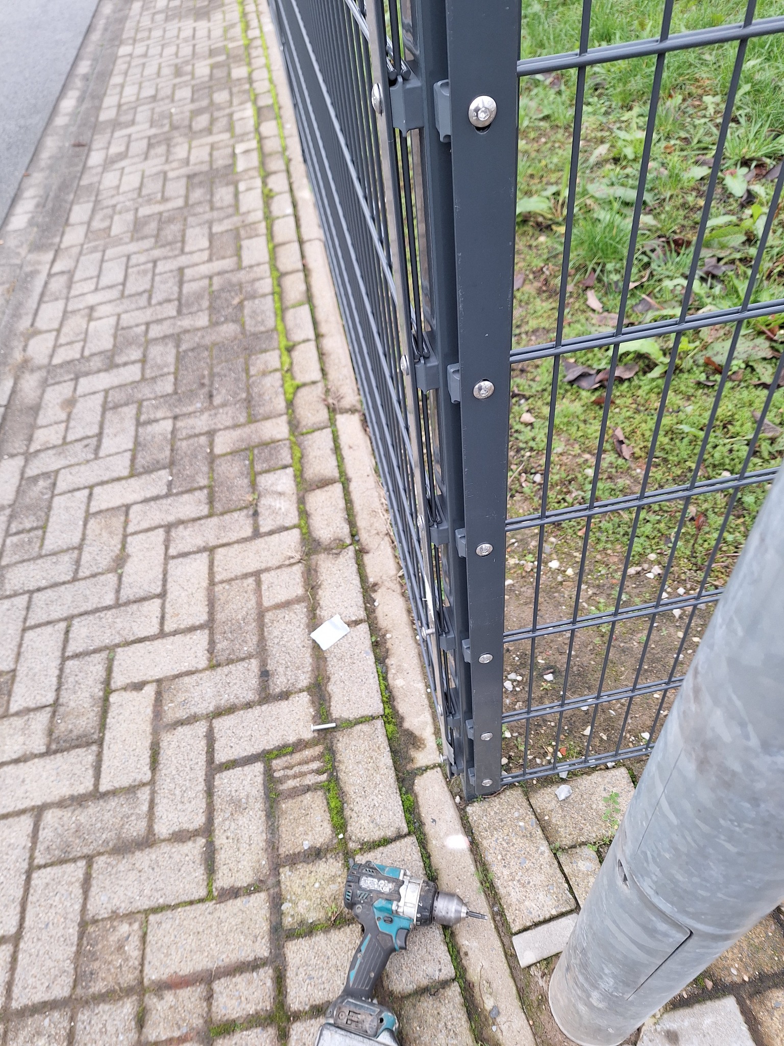 Szary metalowy płot panelowy, częściowo zamontowany na kostce brukowej. W pobliżu leży wkrętarka akumulatorowa. Widoczne ślady mchu między kostkami.