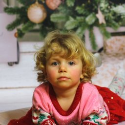 Portret uroczej, kręconowłosej dziewczynki w świątecznym sweterku, leżącej na podłodze z choinką w tle. Ujęcie z bliska, skupione na twarzy dziecka.