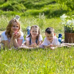 Sesja rodzinna w plenerze: mama z dwójką dzieci leżą na kocu w trawie, obok kosz z kwiatami. Uśmiechnięte twarze, naturalne światło, sielski klimat.
