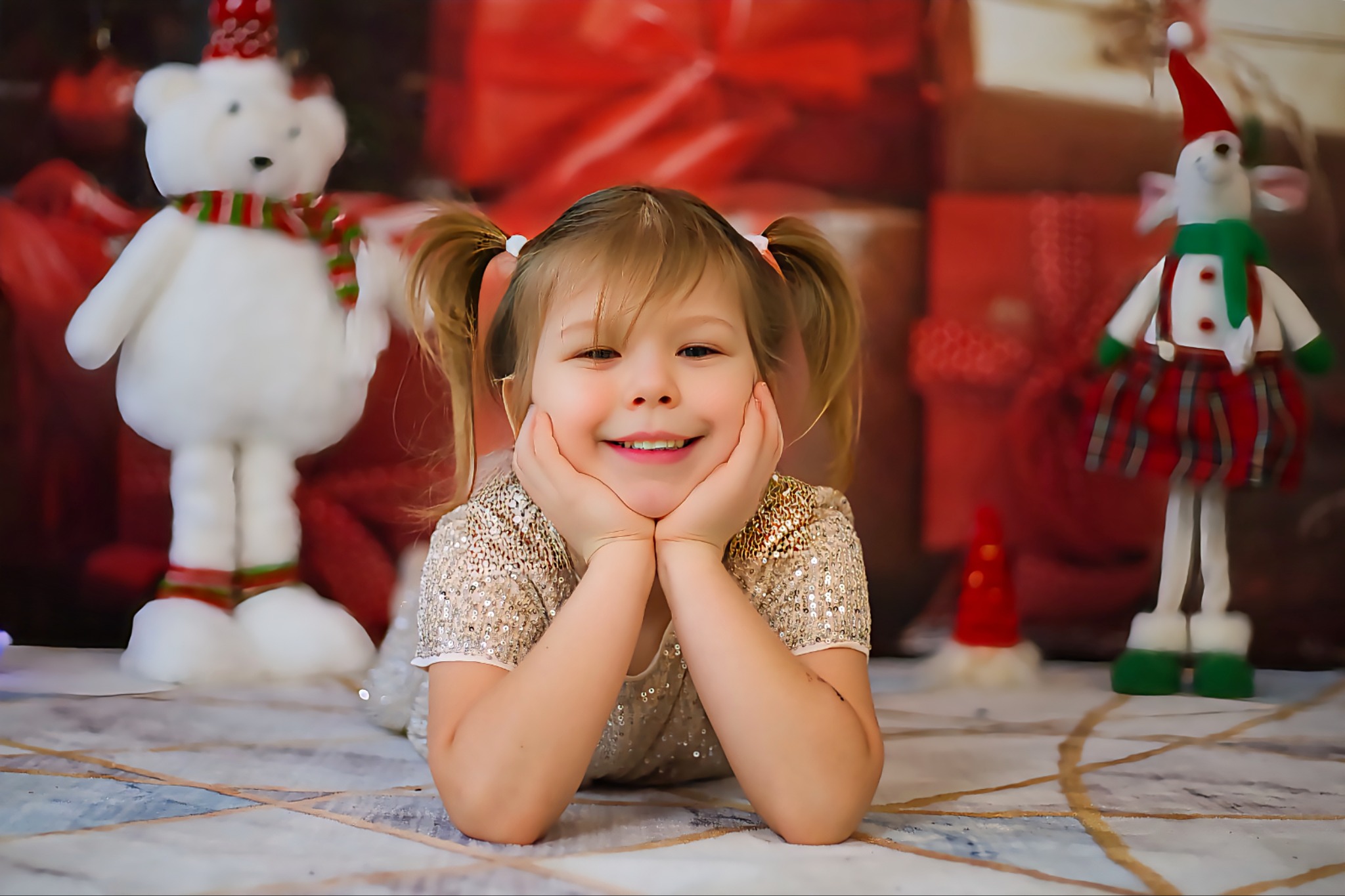 Uśmiechnięta dziewczynka w cekinowej sukience leży na podłodze z zabawkami w świątecznym tle. Dwa kucyki, ręce pod brodą, radosny wyraz twarzy.