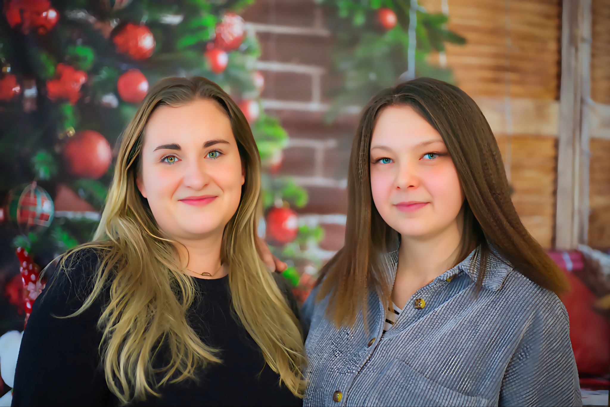 Portret dwóch uśmiechniętych kobiet na tle świątecznej dekoracji z choinką i ceglaną ścianą. Jedna z blond włosami, druga z brązowymi.
