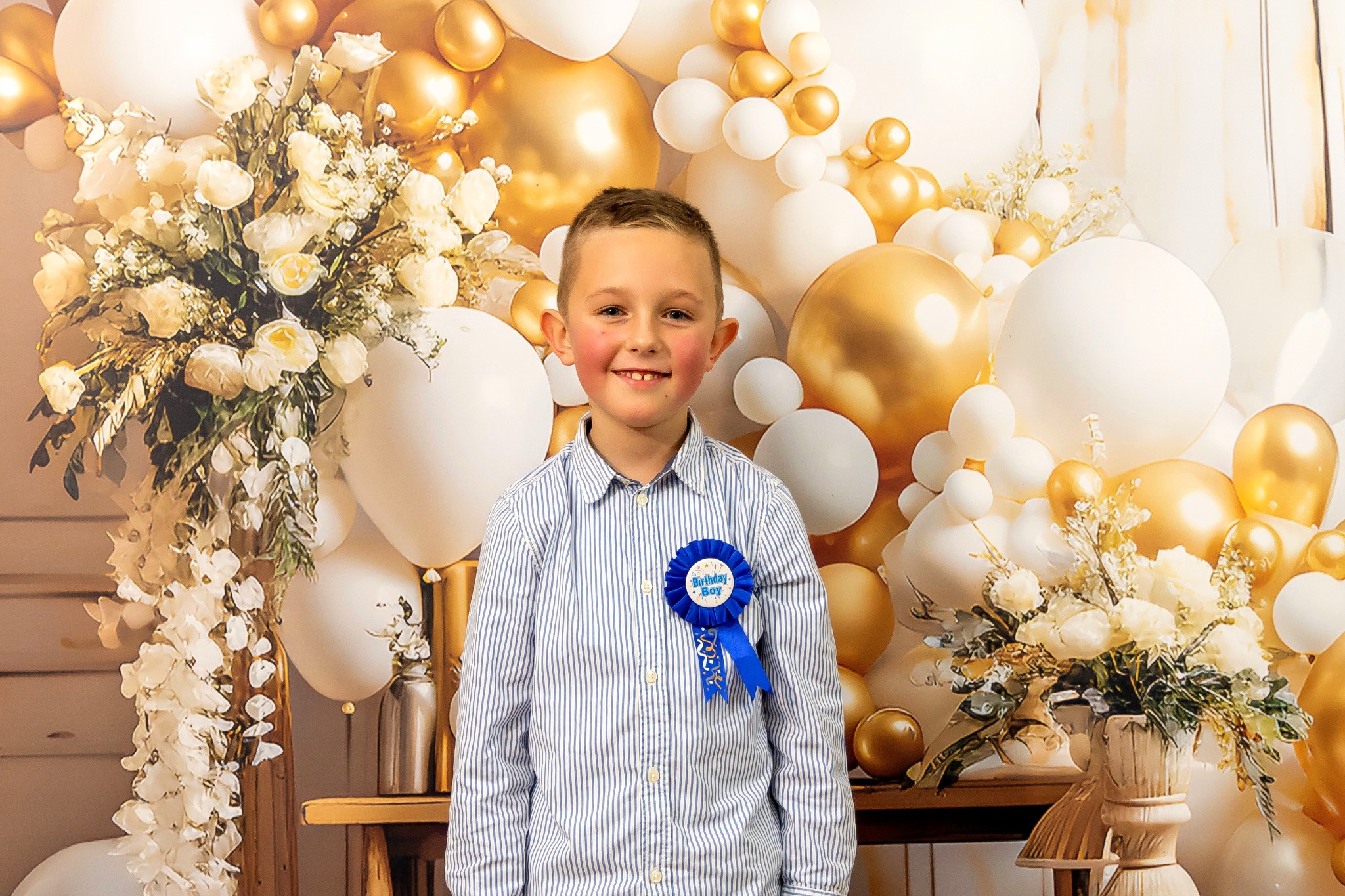 Uśmiechnięty chłopiec z przypinką 'Birthday Boy' na tle dekoracji z białych i złotych balonów oraz kwiatów. Sesja urodzinowa w jasnej tonacji.