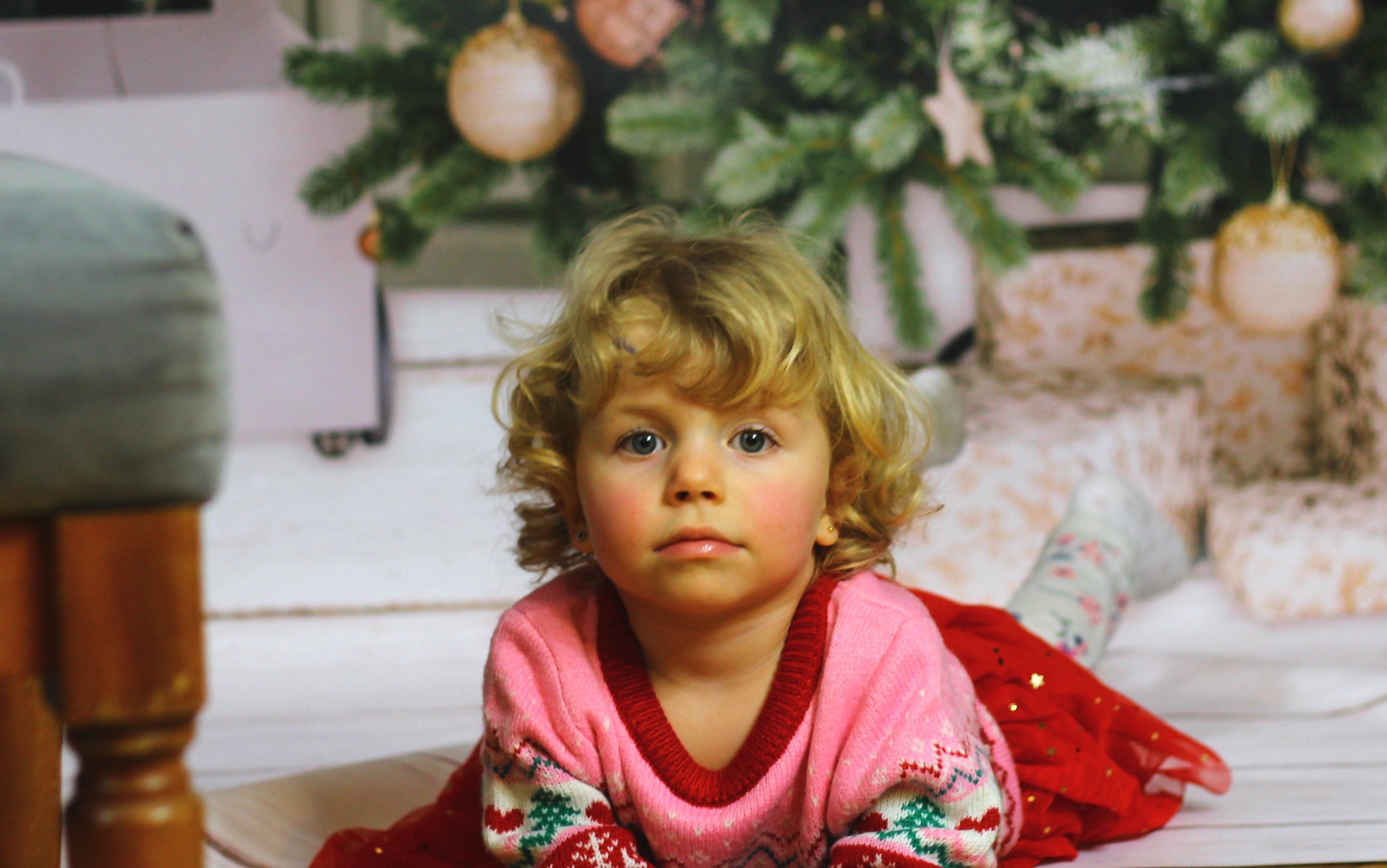 Portret uroczej, kręconowłosej dziewczynki w świątecznym sweterku, leżącej na podłodze z choinką w tle. Ujęcie z bliska, skupione na twarzy dziecka.