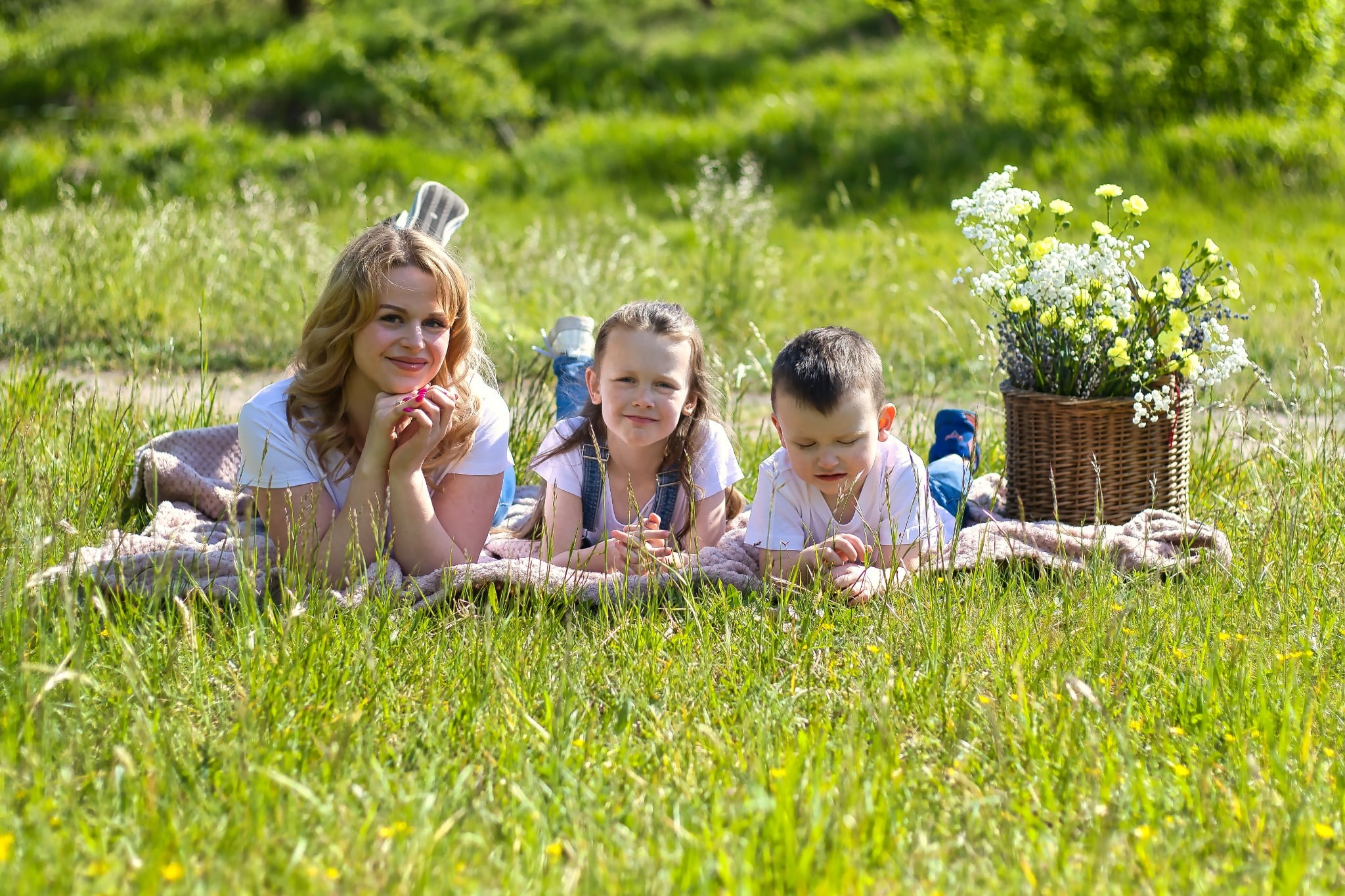 Sesja rodzinna w plenerze: mama z dwójką dzieci leżą na kocu w trawie, obok kosz z kwiatami. Uśmiechnięte twarze, naturalne światło, sielski klimat.