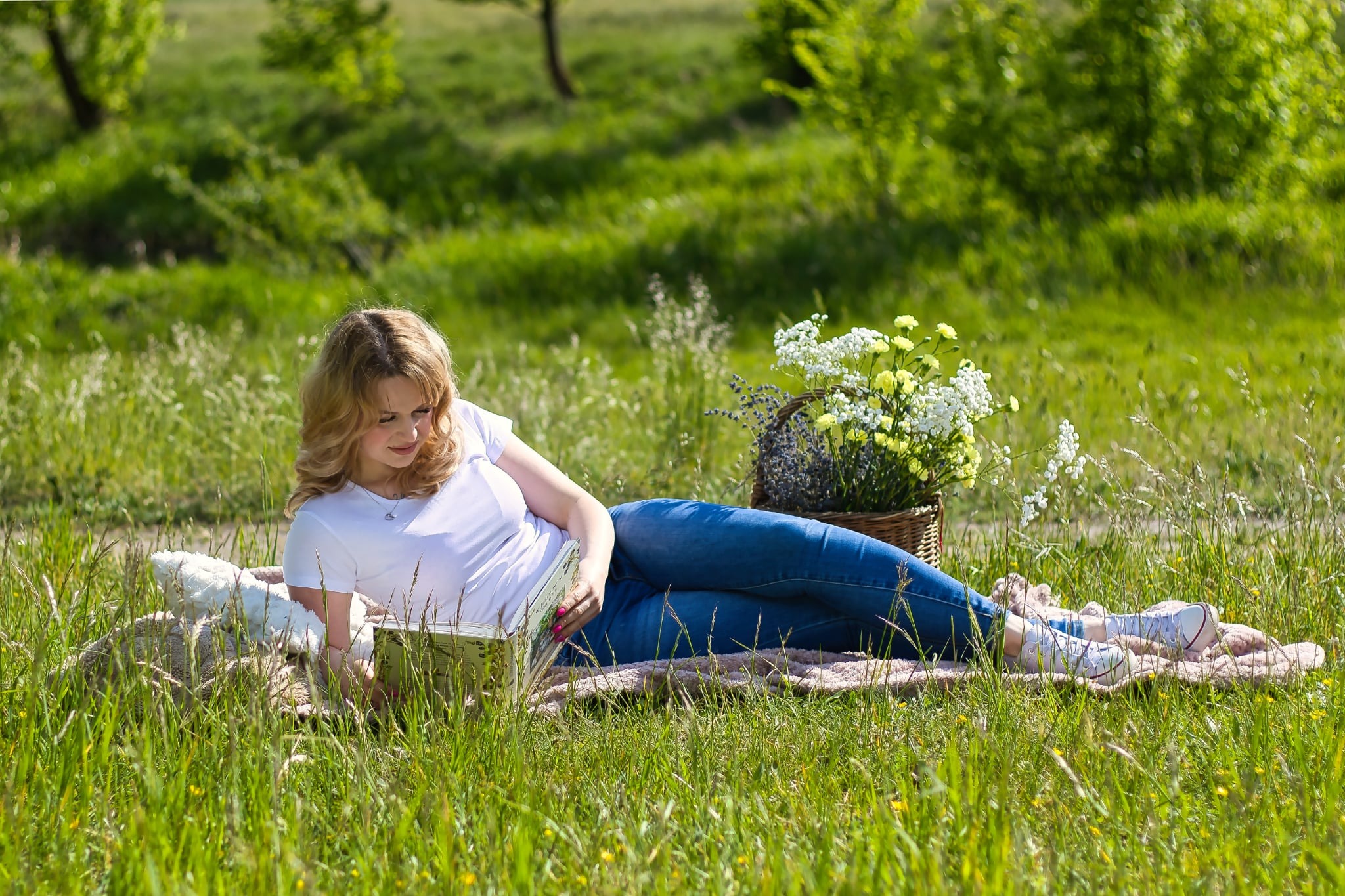 Blondynka w jeansach czyta książkę na kocu w trawie obok koszyka z kwiatami. Słoneczny dzień, relaks na łonie natury. Ujęcie z niskiej perspektywy.
