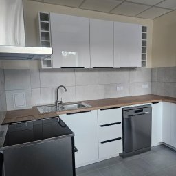 Clean Kropla-Sprzątanie Częstochowa - Nowoczesna, jasna kuchnia po remoncie: białe szafki z czarnymi uchwytami, drewniany blat, szare płytki i podłoga. Widoczny fragment płyty indukcyjnej i zmywarki.