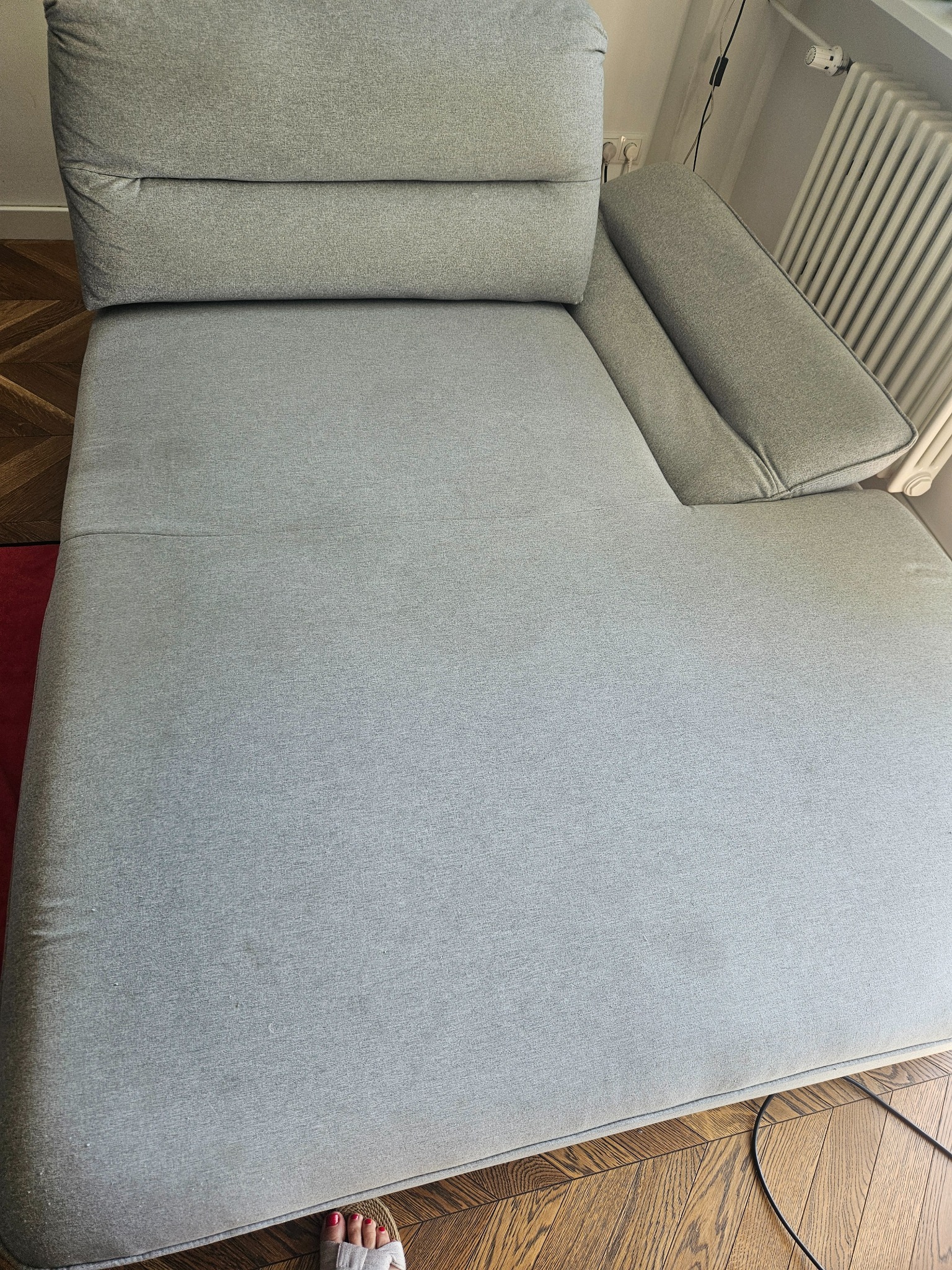 Jasnoszara, modułowa sofa narożna z widocznymi drobnymi zabrudzeniami na siedzisku, ustawiona na parkiecie. Fragment stopy w klapku w rogu kadru.