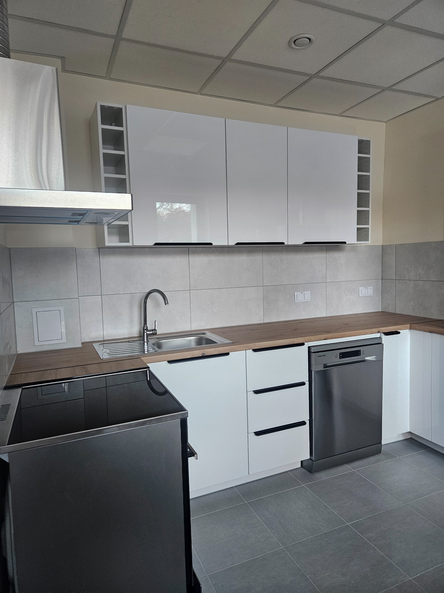 Nowoczesna, jasna kuchnia po remoncie: białe szafki z czarnymi uchwytami, drewniany blat, szare płytki i podłoga. Widoczny fragment płyty indukcyjnej i zmywarki.