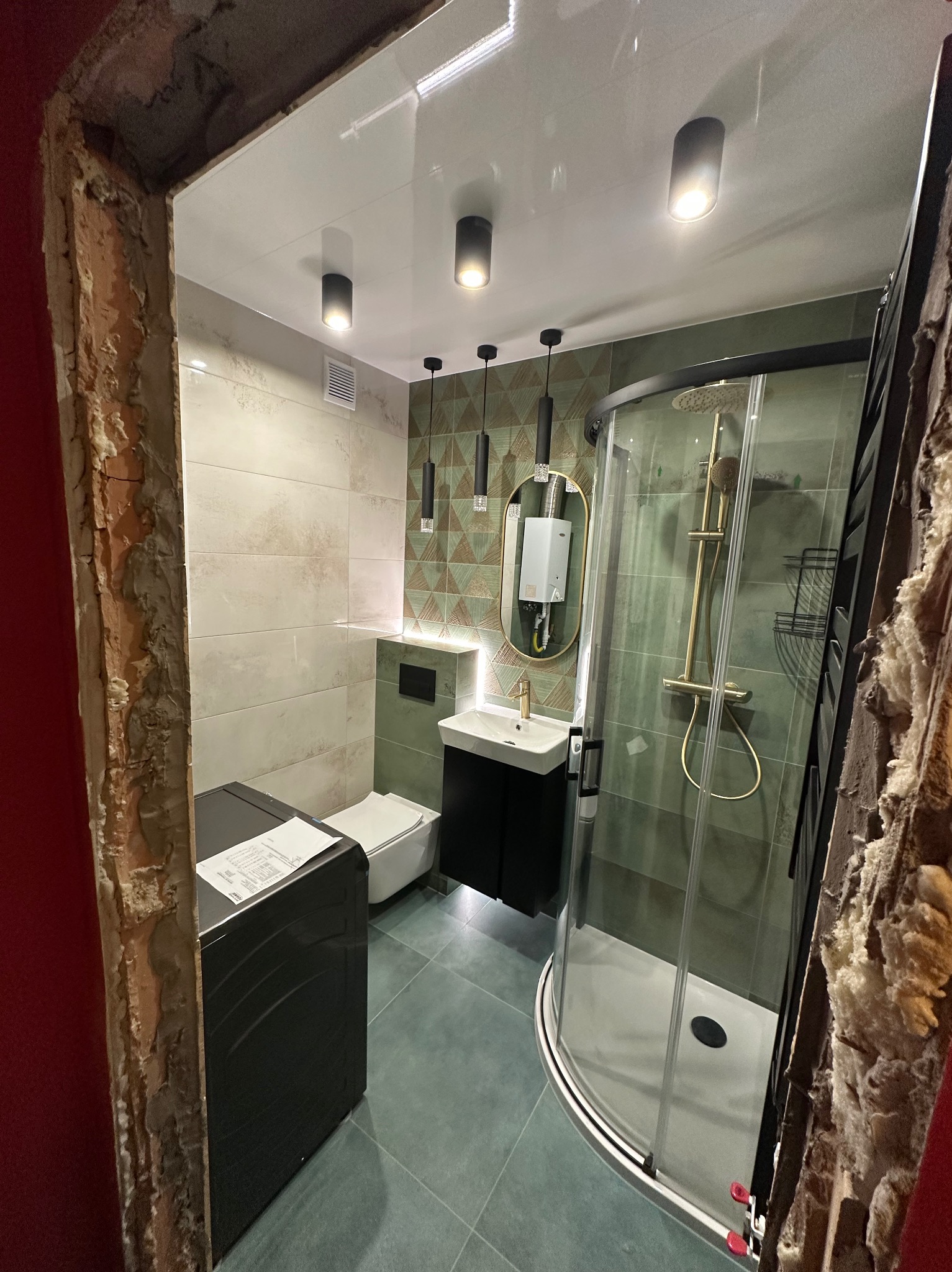 Nowoczesna łazienka po remoncie w Rudnikach: kabina prysznicowa, umywalka z szafką, toaleta i pralka. Ściany wyłożone płytkami w odcieniach beżu i zieleni, designerskie oświetlenie.