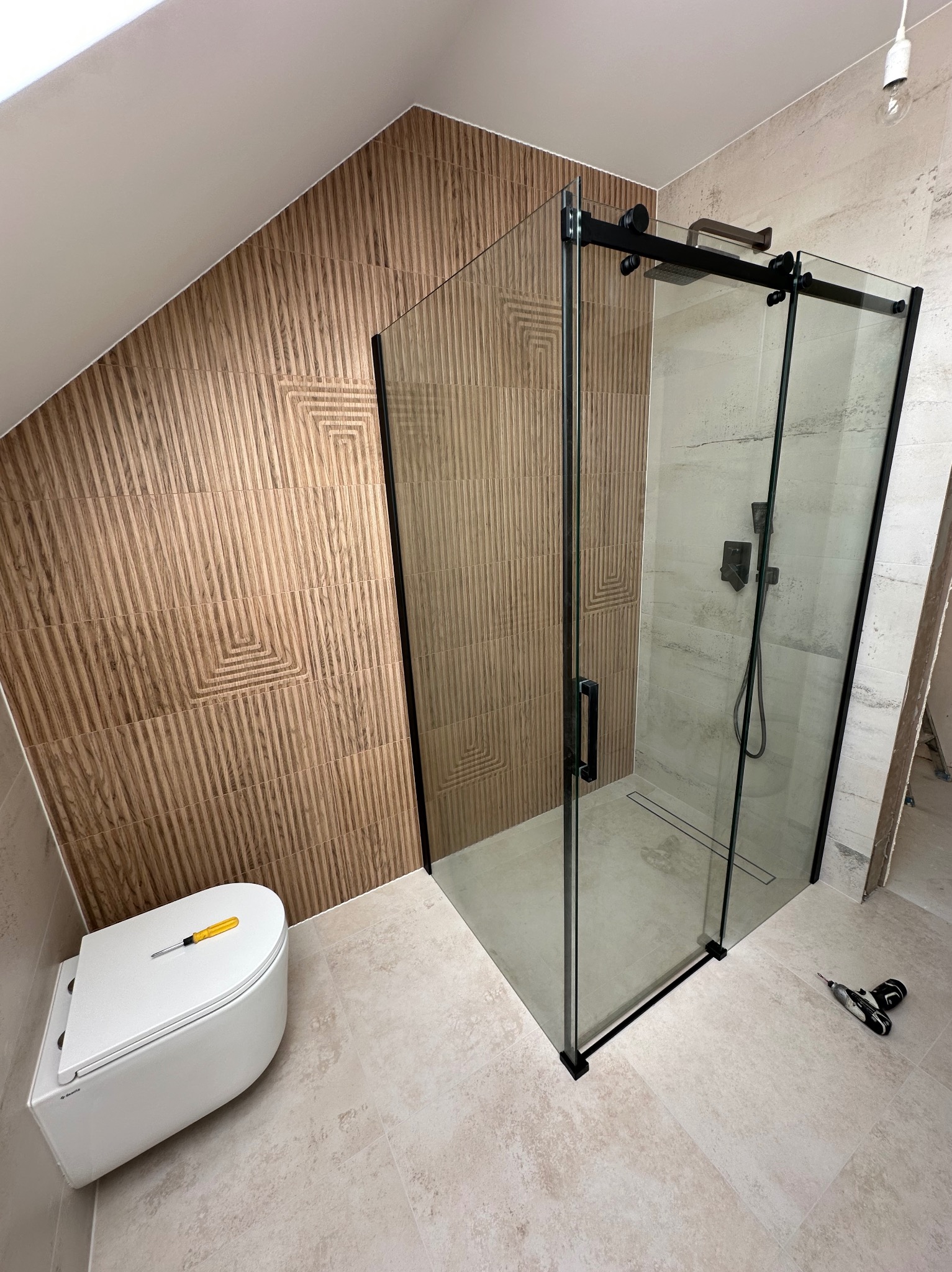 Wykończona łazienka na poddaszu: kabina prysznicowa z czarnymi profilami, toaleta z żółtym śrubokrętem, płytki imitujące drewno na skośnej ścianie.