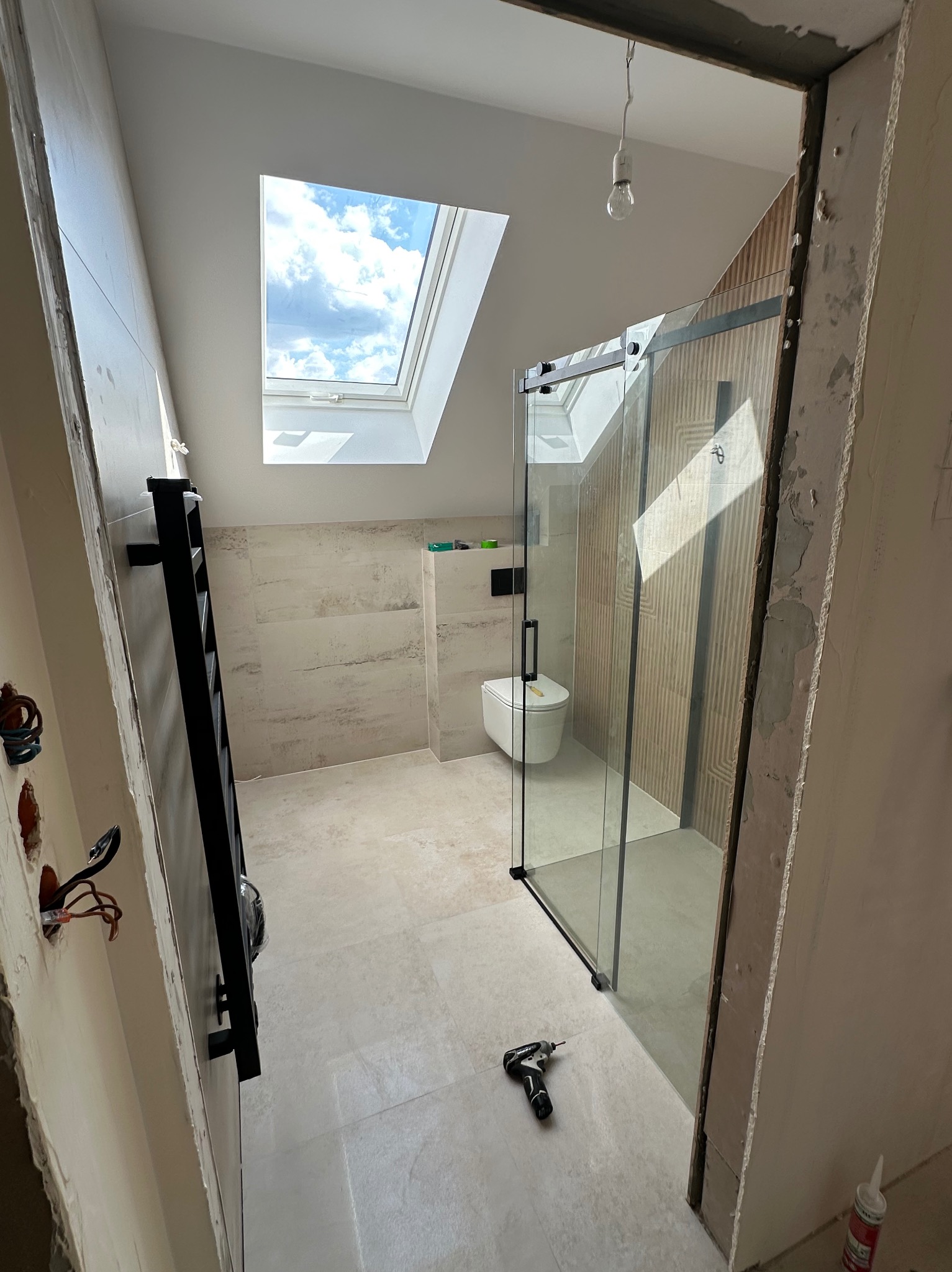 Wykończona łazienka na poddaszu z kabiną prysznicową typu walk-in, grzejnikiem drabinkowym i oknem dachowym. W tle widoczna toaleta. Na podłodze leży wkrętarka.