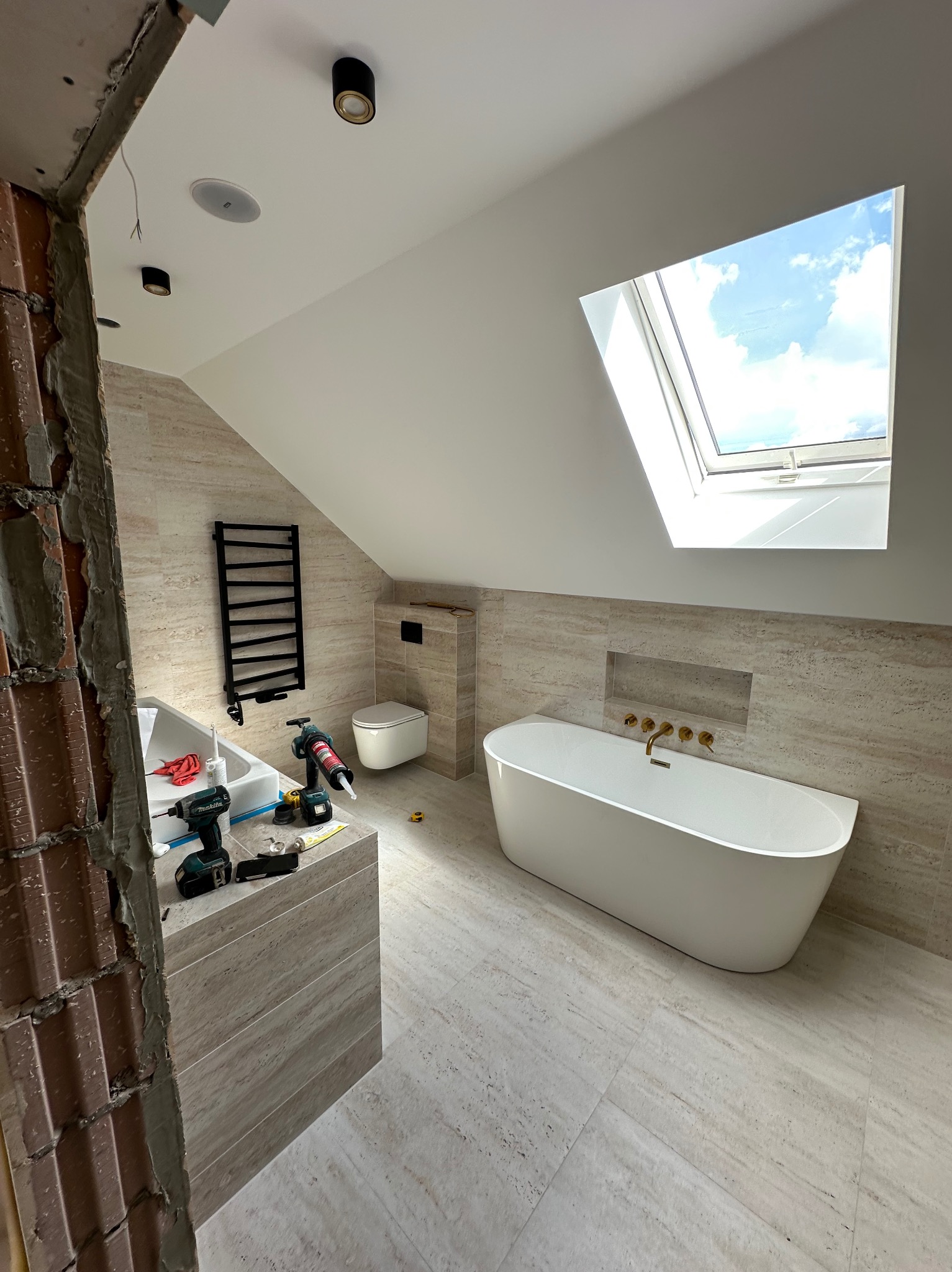 Remont łazienki na poddaszu: wanna wolnostojąca, płytki imitujące kamień, okno dachowe. Widoczne narzędzia i fragment ściany w stanie surowym.