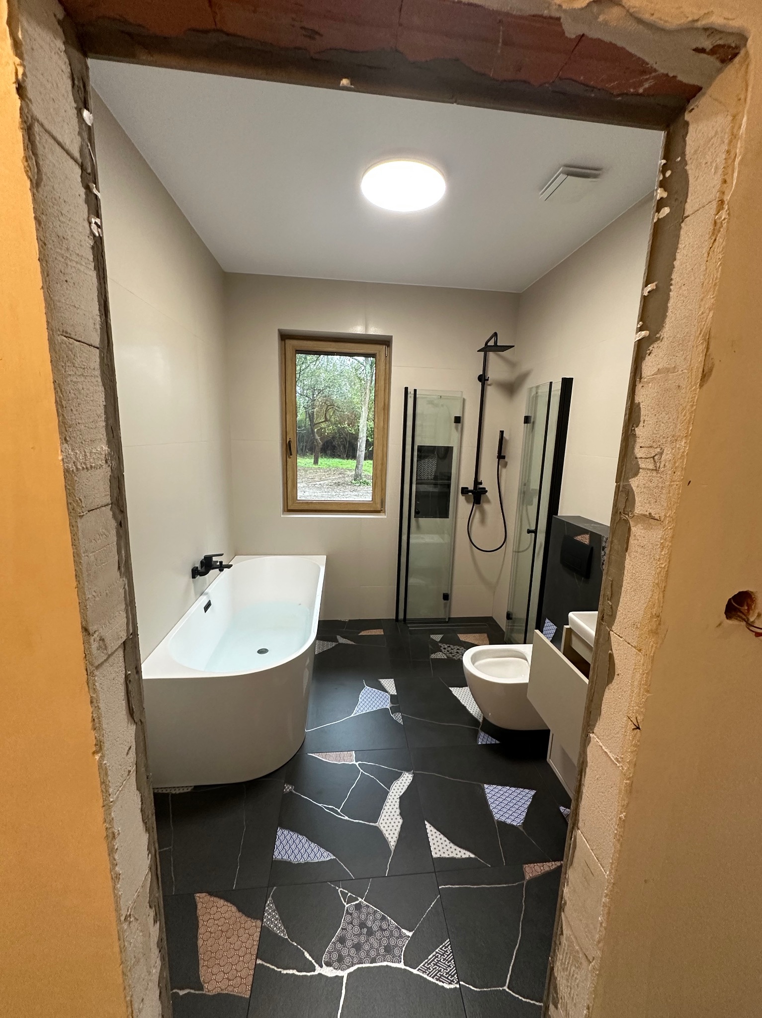 Nowoczesna łazienka z czarno-białą mozaiką na podłodze, białą wanną i kabiną prysznicową z czarnymi ramami, widziana przez surowy otwór drzwiowy.